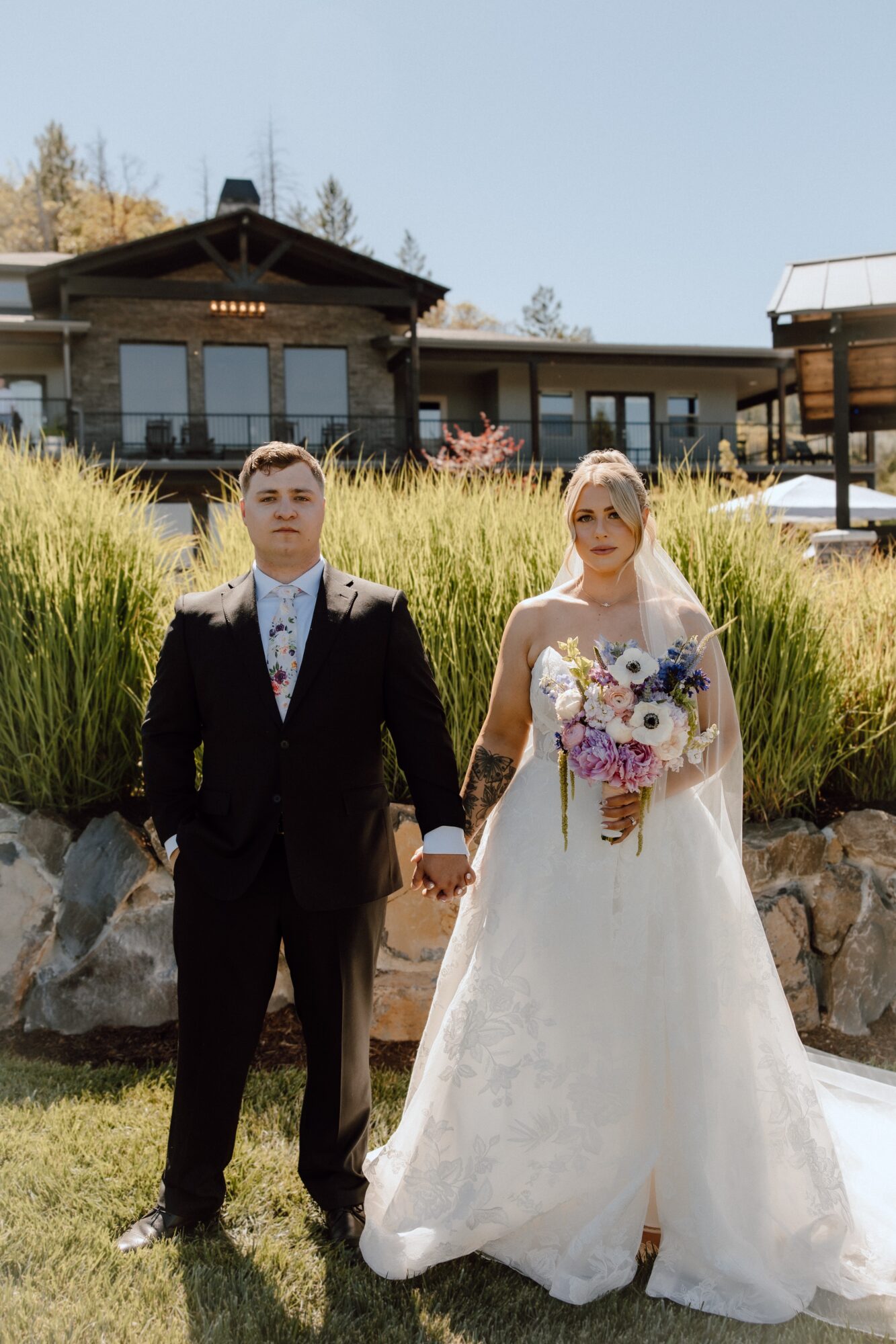 A bride and groom holding hands outdoors, with a building and landscaped garden in the background.