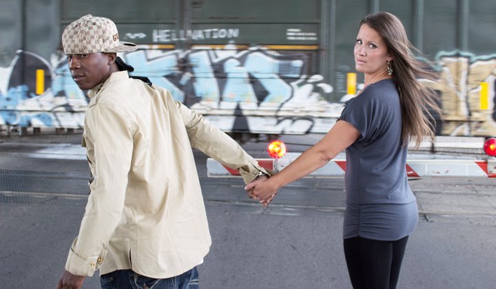 Two women holding hands walk on a city street with graffiti-covered walls in the background.