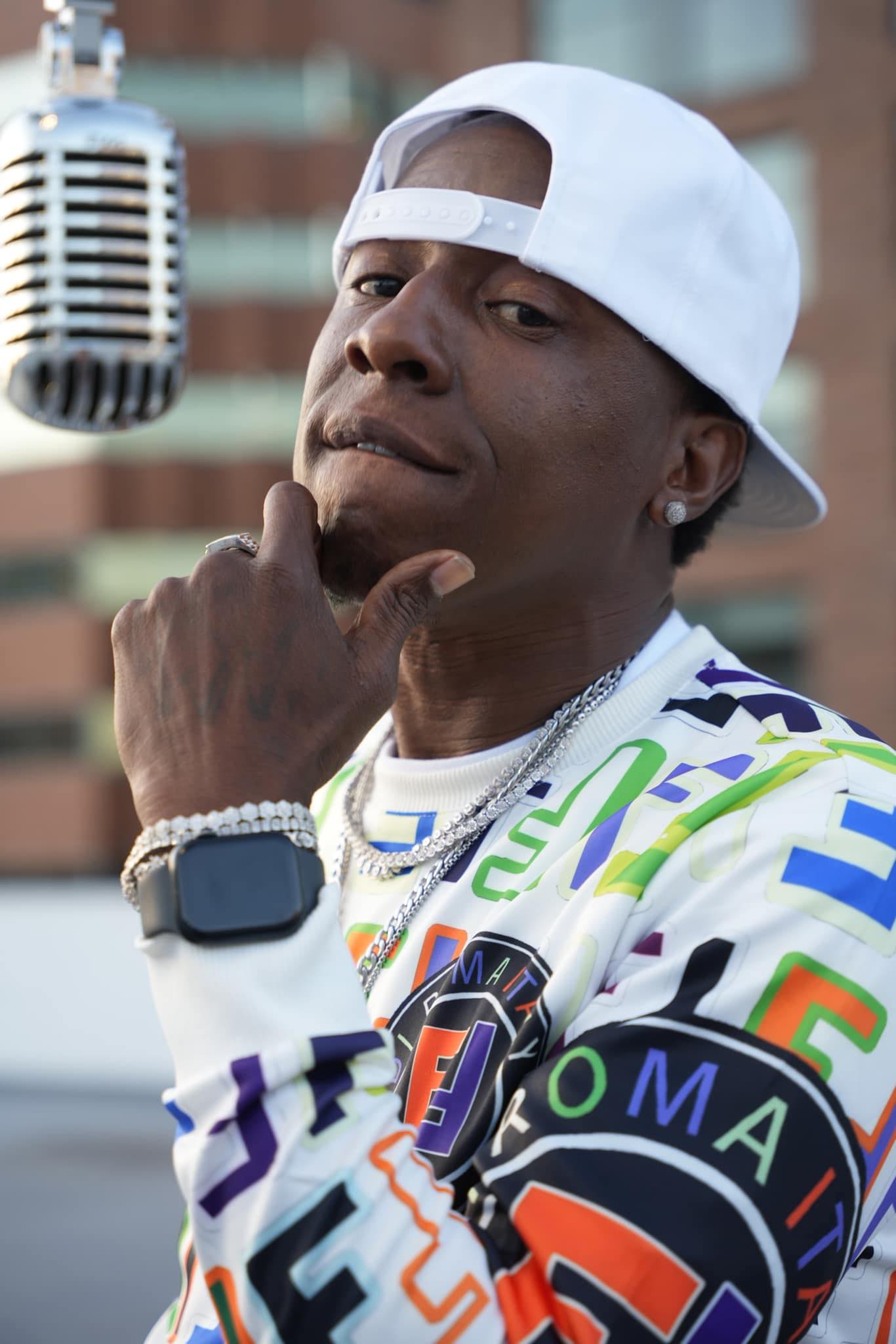 Man wearing a white cap backwards, looking at camera with hand on chin, outdoors with microphone nearby, city buildings in background.