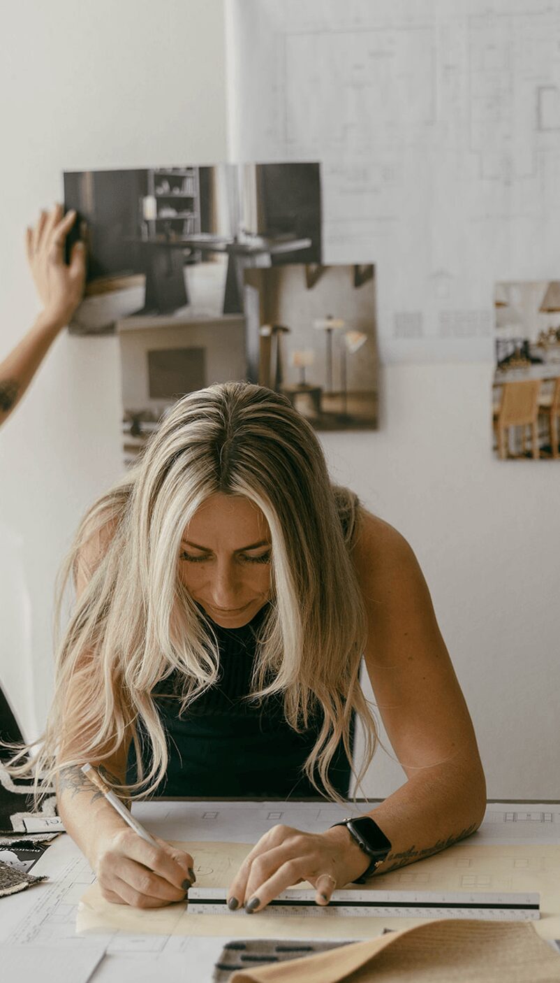 Woman with long blonde hair writing on paper at desk, background with architectural plans and photos