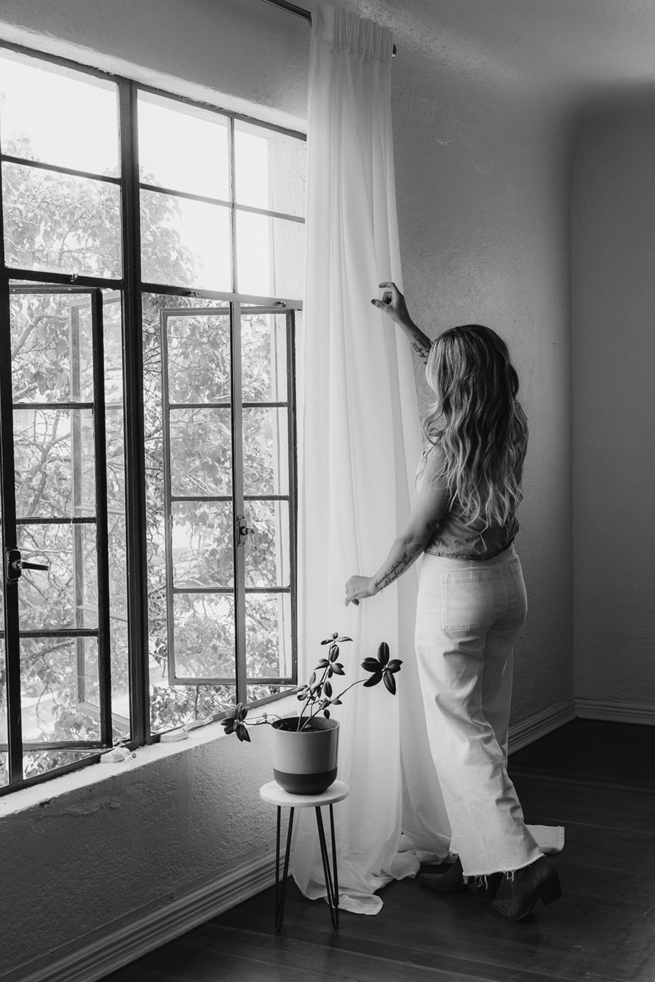 Woman adjusting curtains by a large window with a potted plant on a stand, natural light coming in, black and white photo.