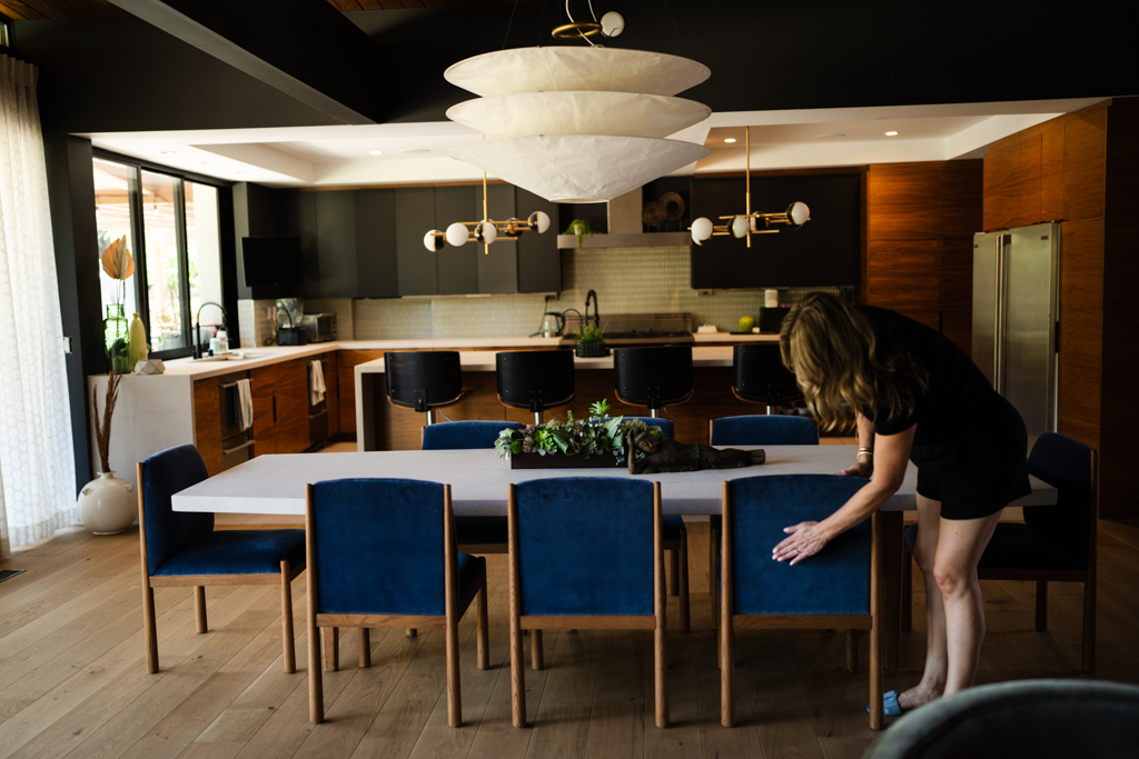 Woman arranging chairs around a dining table in a modern kitchen and dining area, with large windows and contemporary lighting fixtures.