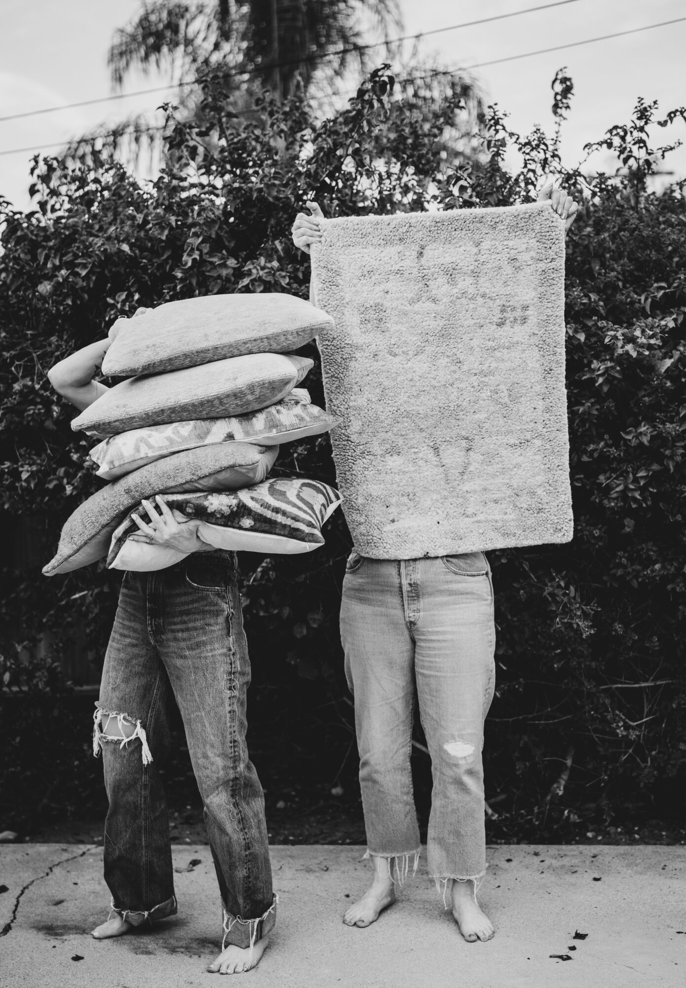 Person with a towel on their head holding a large stack of pillows, next to a person with a towel covering their head, standing outdoors.