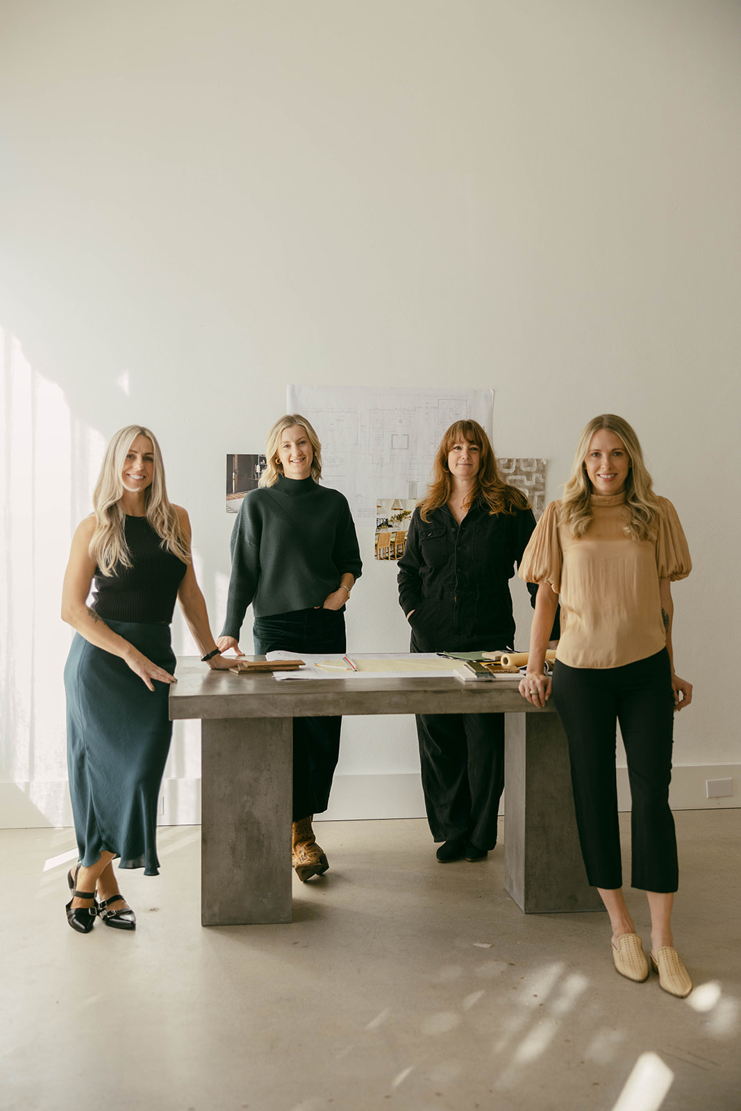 Four women stand behind a table in a bright room with white walls and large windows.