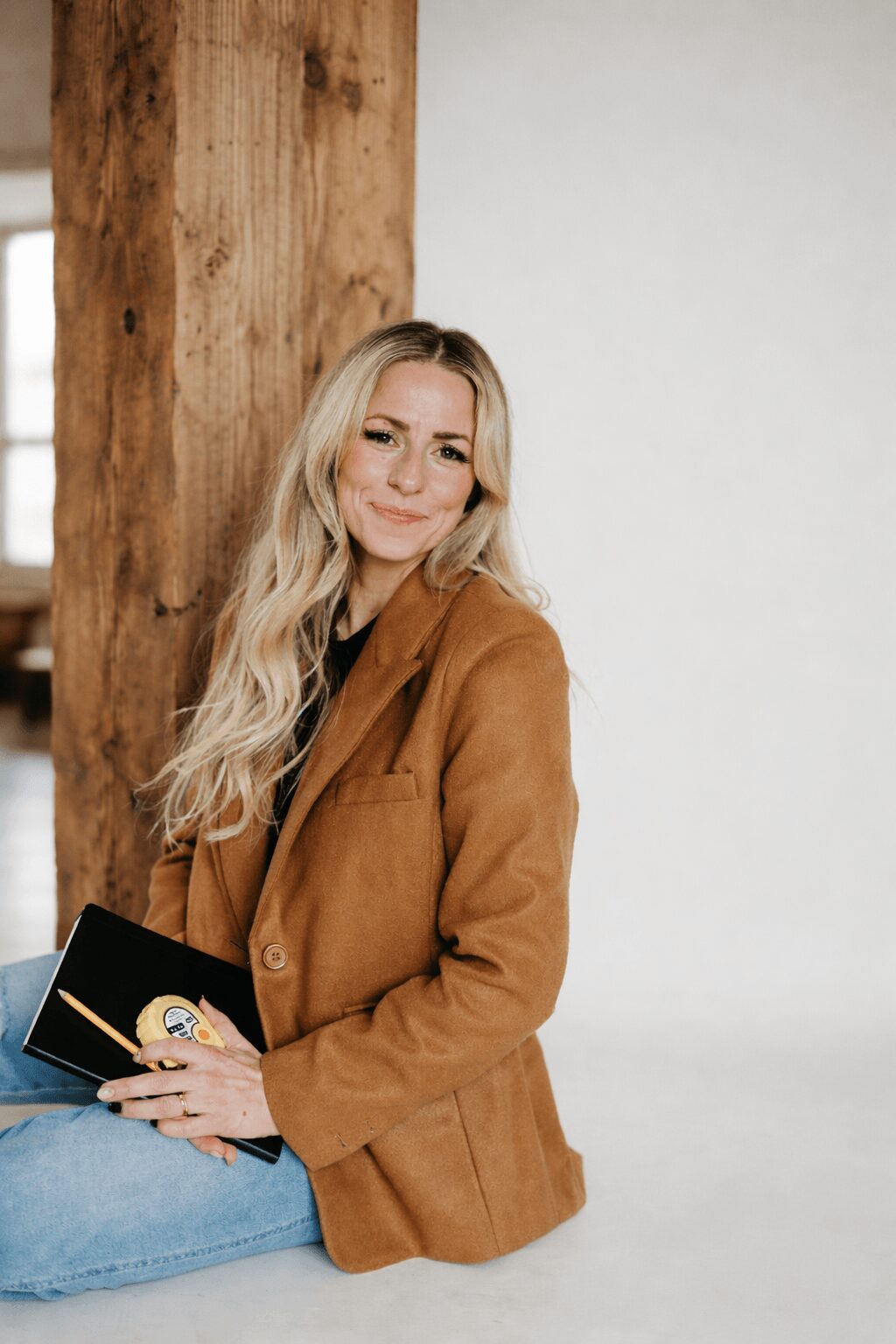 Woman with long blonde hair wearing a brown coat, holding a book, sitting against a wooden post and white wall.