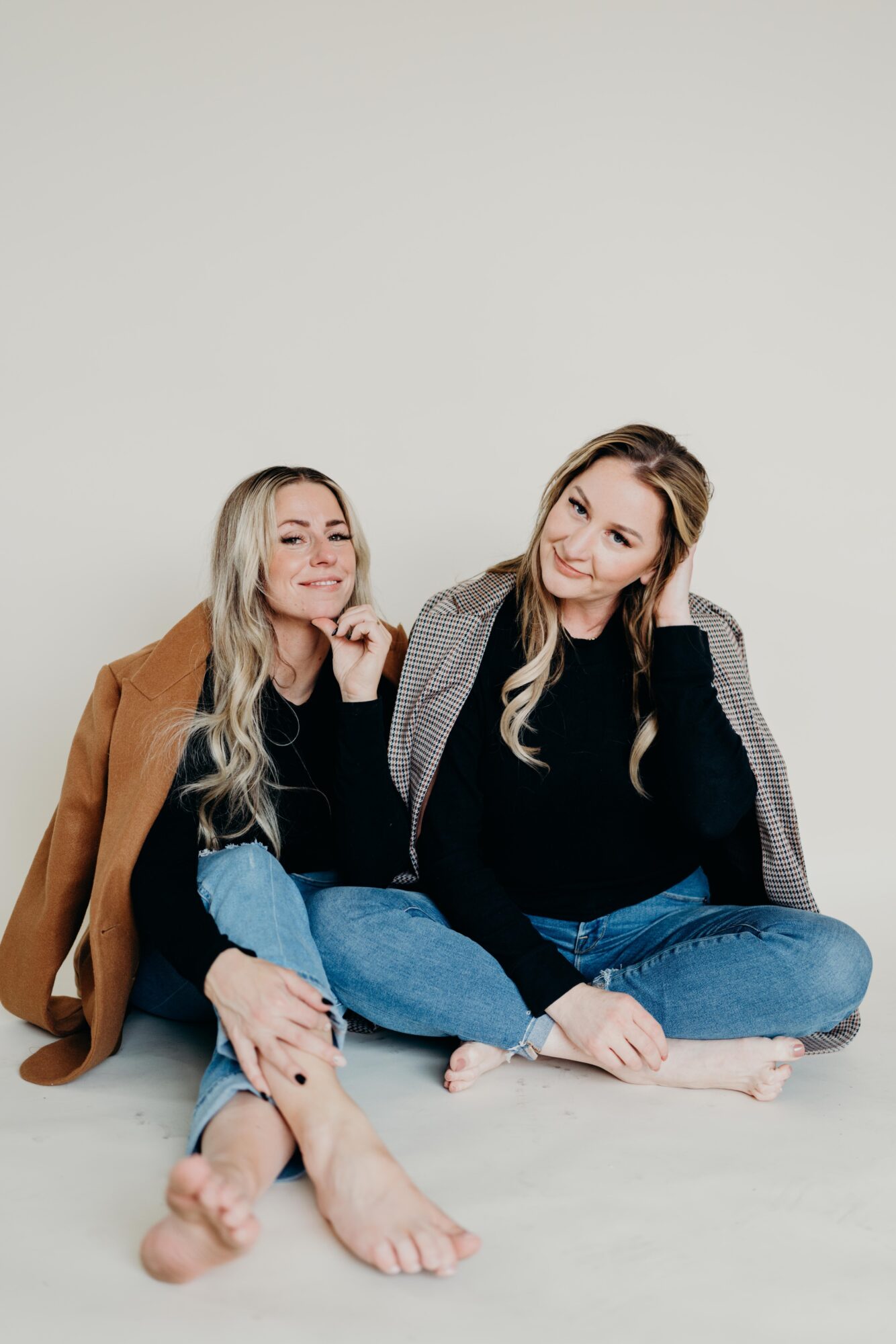 Two women sit on the floor against a plain background, smiling and leaning towards each other, one with a coat, the other with a blazer.