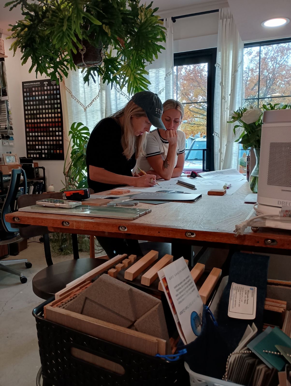 Two women sit at a table, looking at papers, in a room with large windows and plants.
