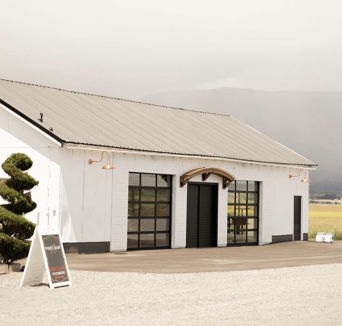 White building with black window frames and a wooden door, outdoor setting with a tree and a signboard, cloudy sky.
