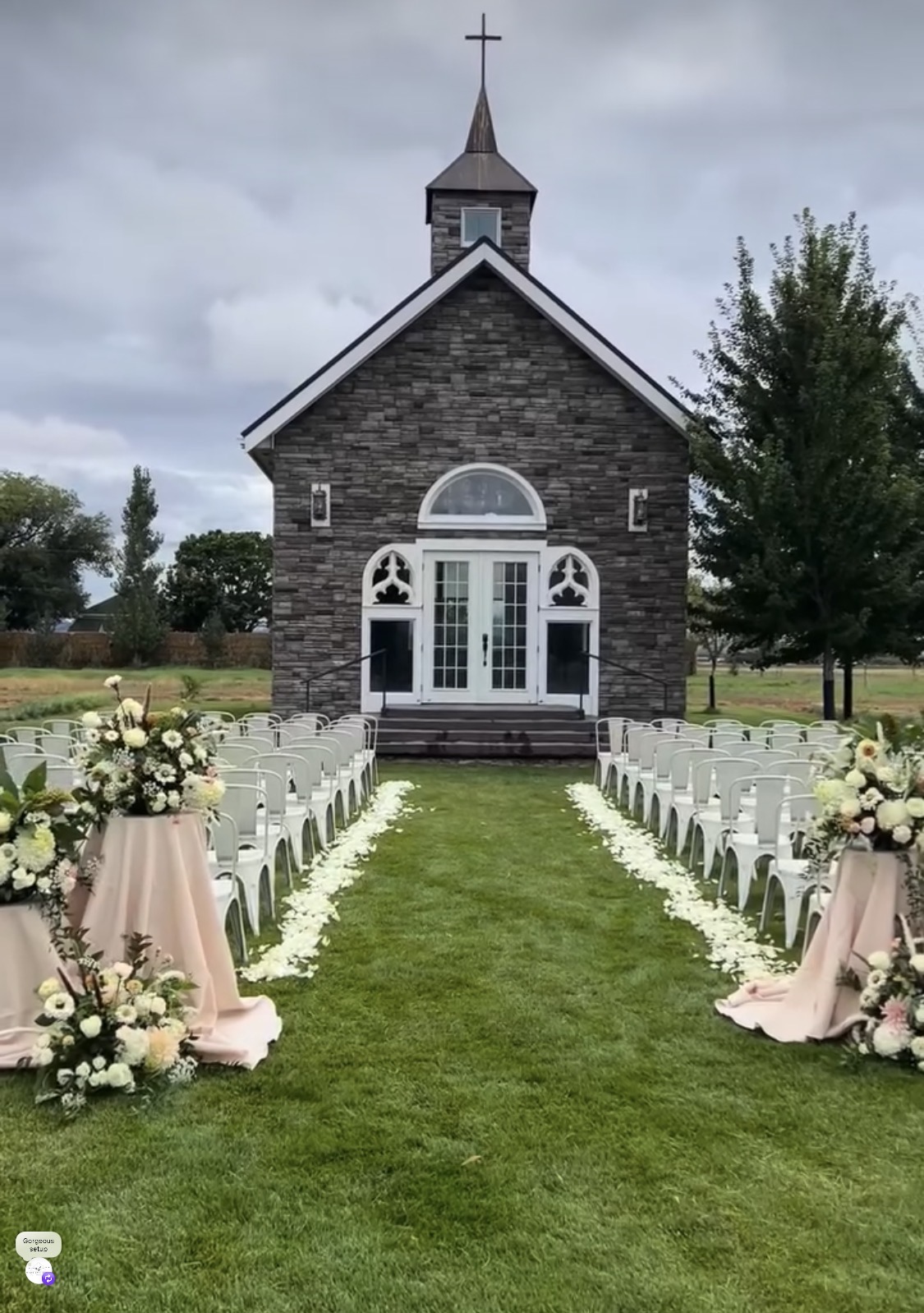 Small stone church with white doors and windows, decorated for outdoor wedding, surrounded by floral arrangements and chairs, cloudy sky.