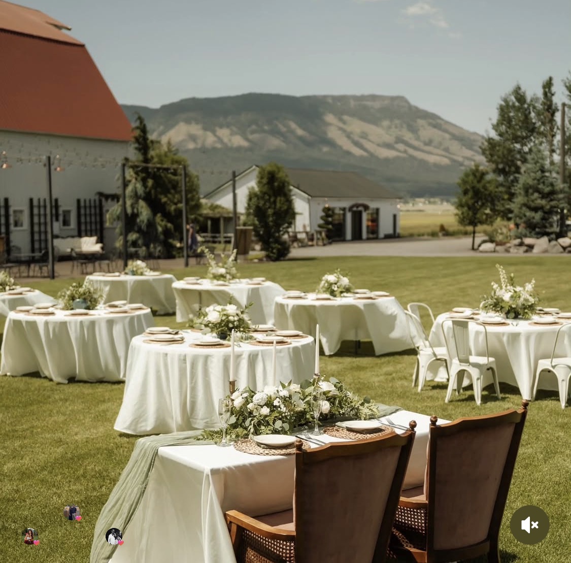 Outdoor event setup with round tables covered in white cloths, floral centerpieces, chairs, and a scenic mountain view in the background.