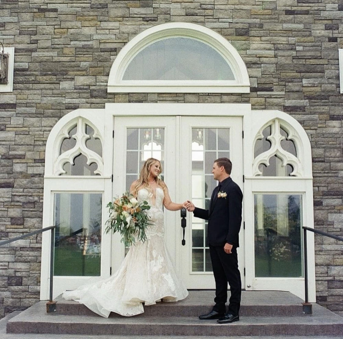 Bride and groom holding hands in front of a stone building with arched windows and white doors.