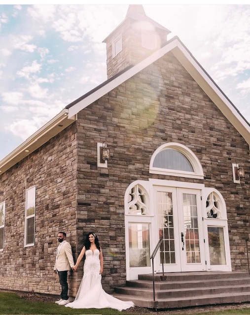 A couple stands outside a stone church with arched windows and steps, under a partly cloudy sky.