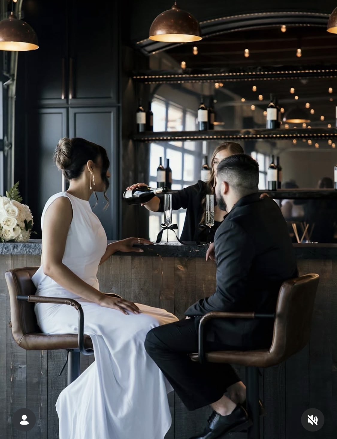 Woman in white dress sitting at bar, talking to man in black suit, in a dimly lit modern bar setting.