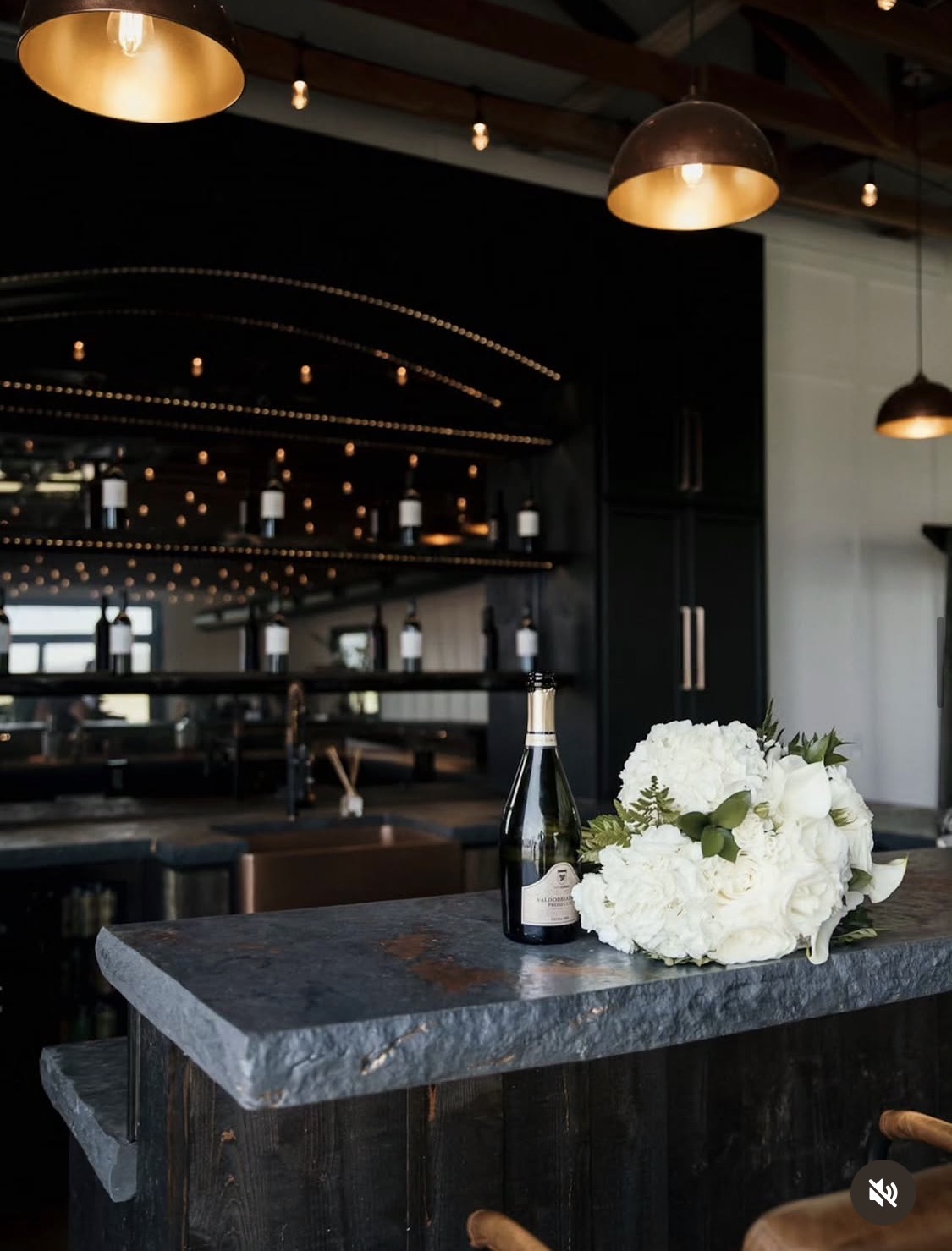 A dark countertop with a bottle of champagne and a bouquet of white flowers, in a modern restaurant interior with hanging lights.