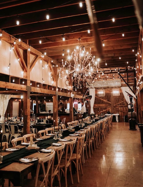 Long dining table set with plates, glasses, and napkins in a rustic, warmly lit venue with string lights and a chandelier.