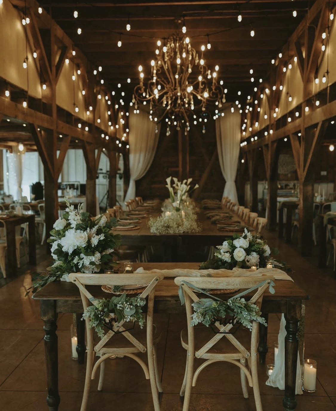 Elegant dining area with a long table, floral centerpieces, chandelier, string lights, and wooden beams, set for a celebration.