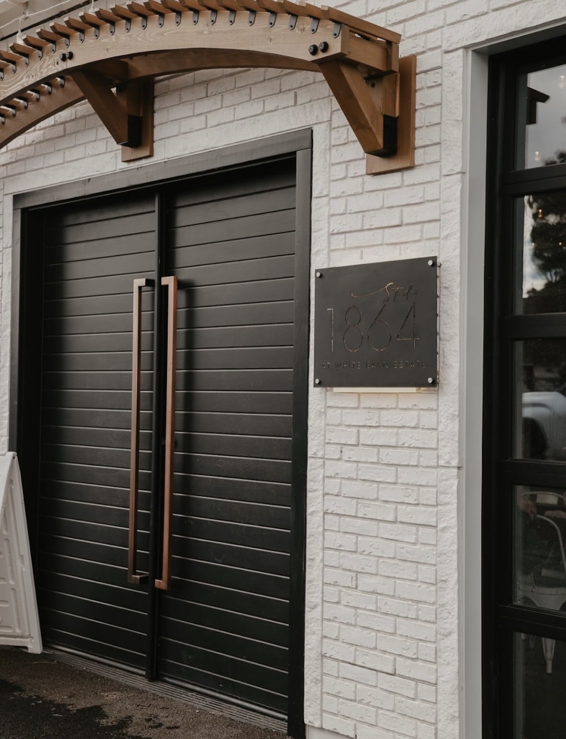 Black sliding door with vertical handles on a white brick building exterior, with a metal plaque and a window nearby.