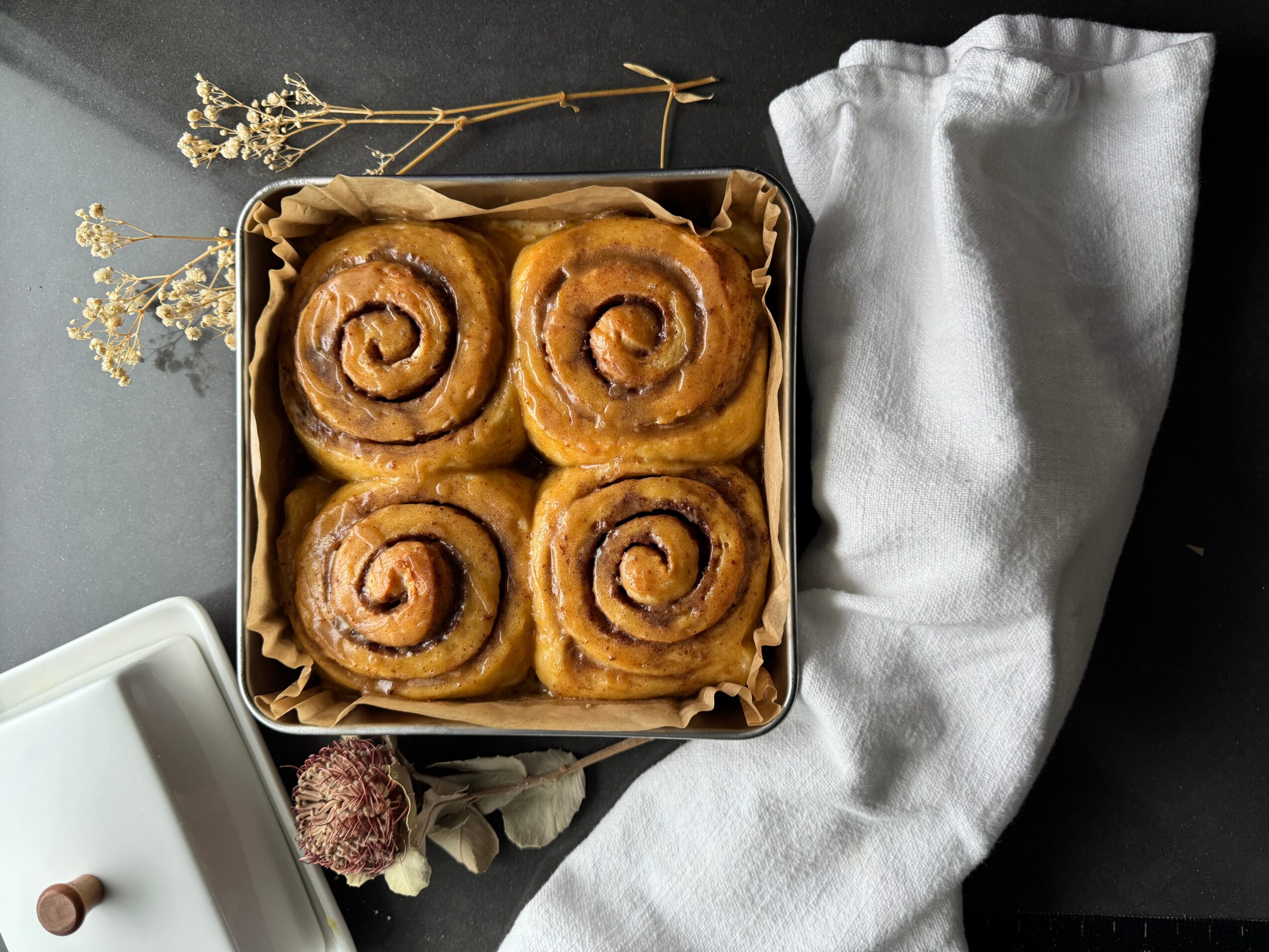 Four cinnamon rolls in a box, with a white cloth and dried flowers nearby, on a dark surface.