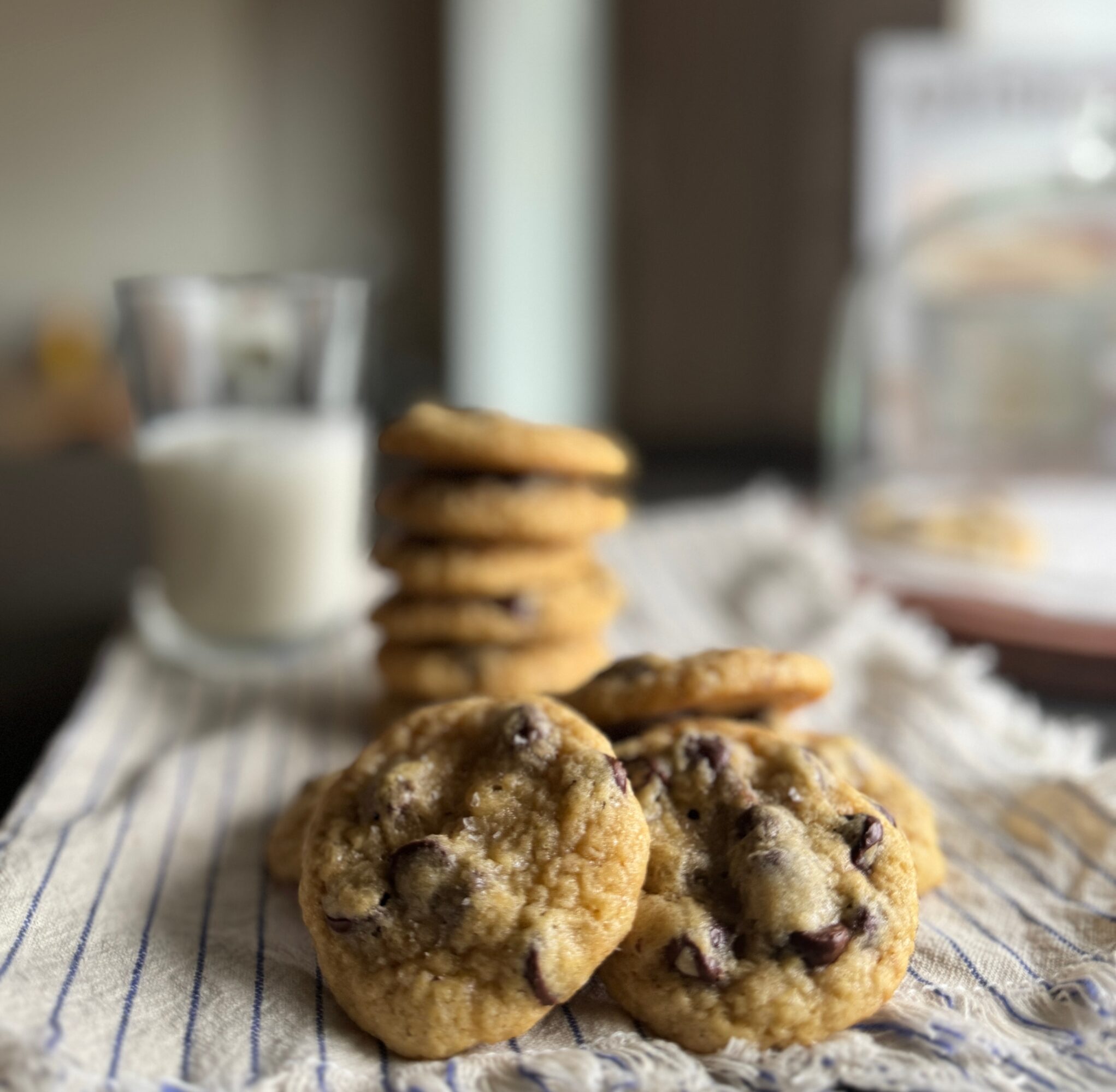 Chocolate chip cookies in front, stack of cookies and a glass of milk in background, on striped cloth.