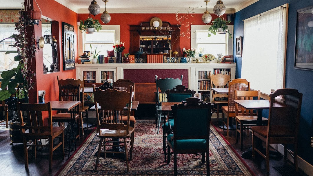 Cozy dining area with colorful walls, wooden chairs, tables, and a bookshelf, decorated with plants and artwork.