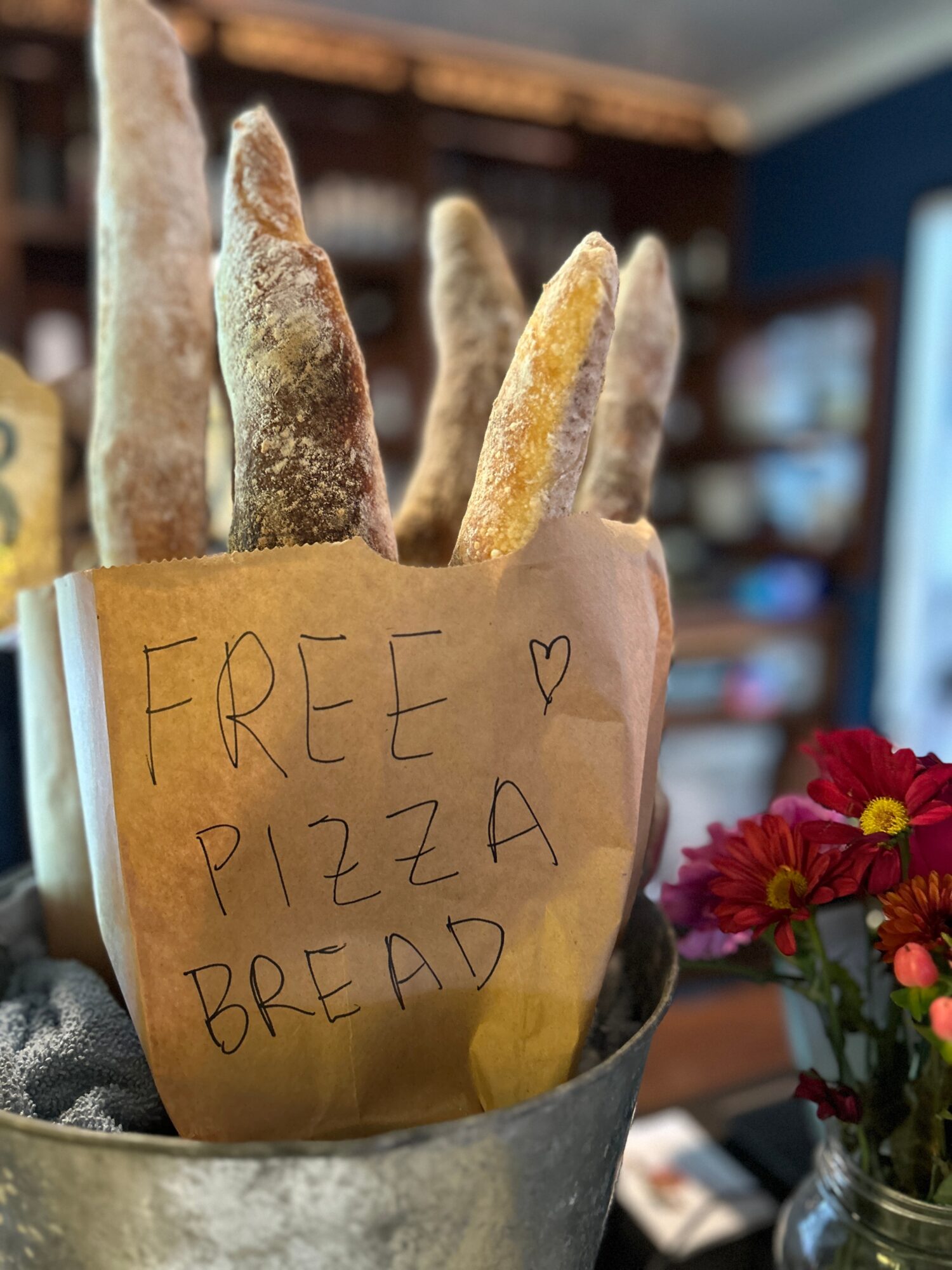 Paper bag with handwritten sign offering free pizza bread, surrounded by breadsticks, next to a vase of pink flowers.