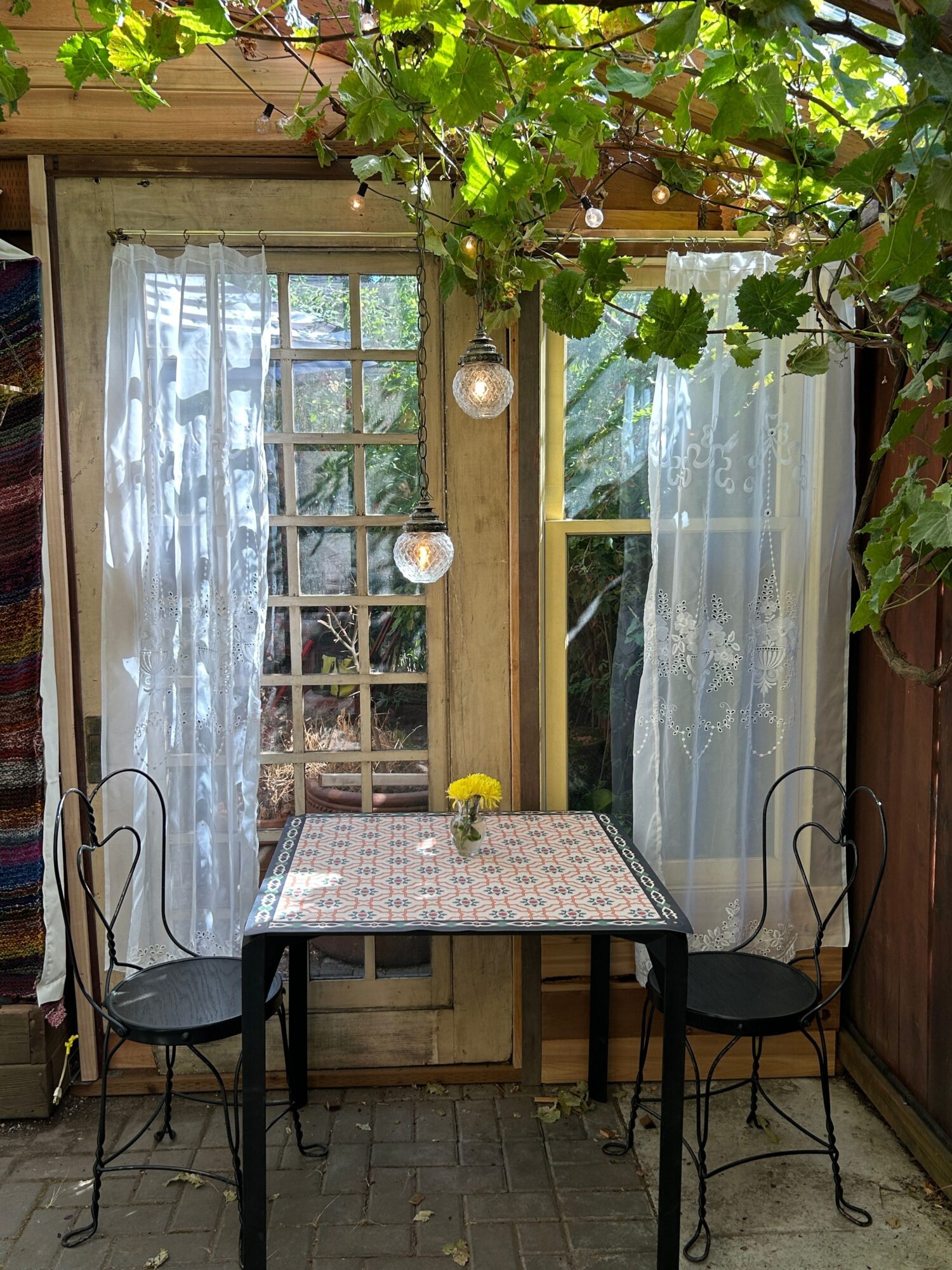 Outdoor patio with a table, chairs, and a windowed door, surrounded by greenery and sunlight.