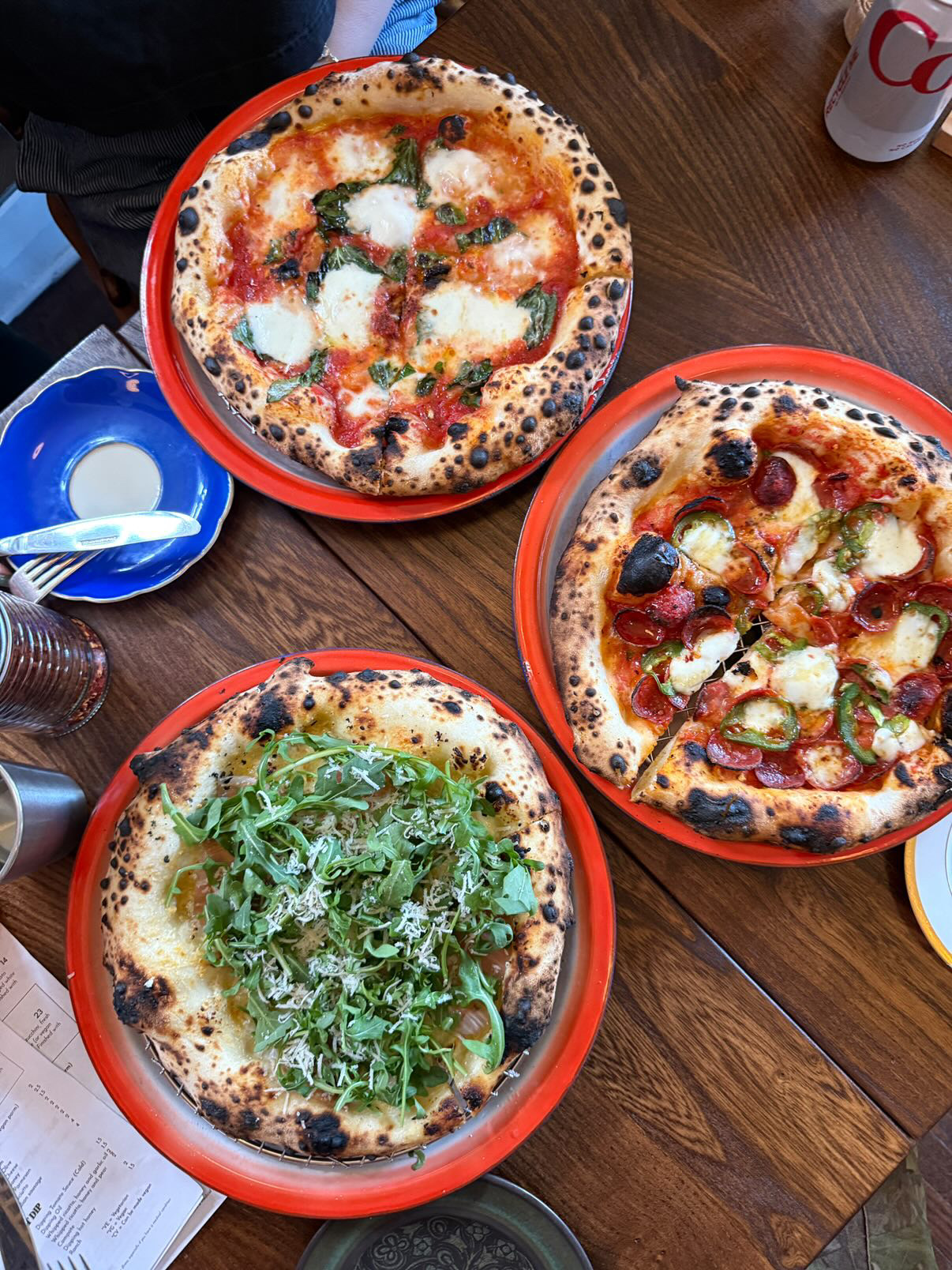 Three pizzas on a wooden table, one with basil, two with various toppings, and a blue plate with a spoon.