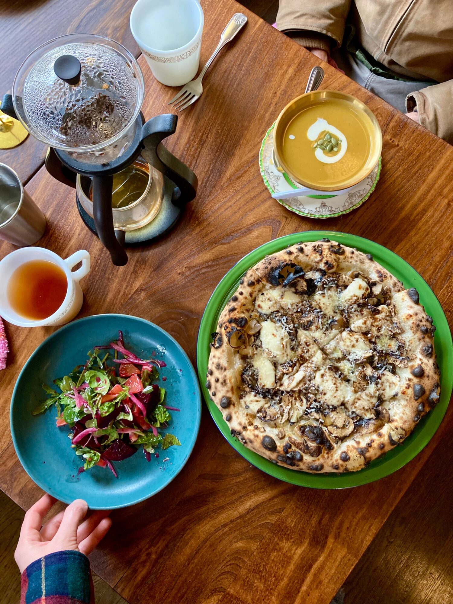Table with a pizza, salad, tea, and drinks, viewed from above, with a person's hand holding a blue plate.