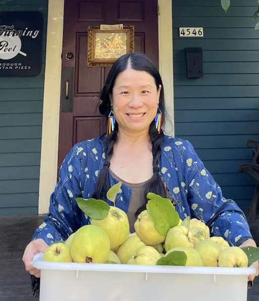 Woman with long dark hair and colorful earrings smiling behind a tray of green apples outside a building.