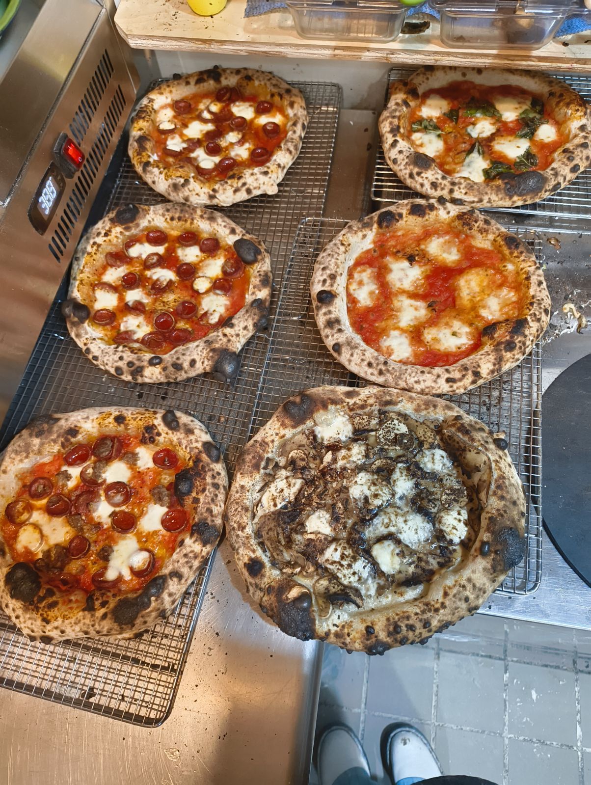 Six pizzas on cooling racks, five with tomato sauce and cheese, one with dark toppings, in a kitchen oven area.