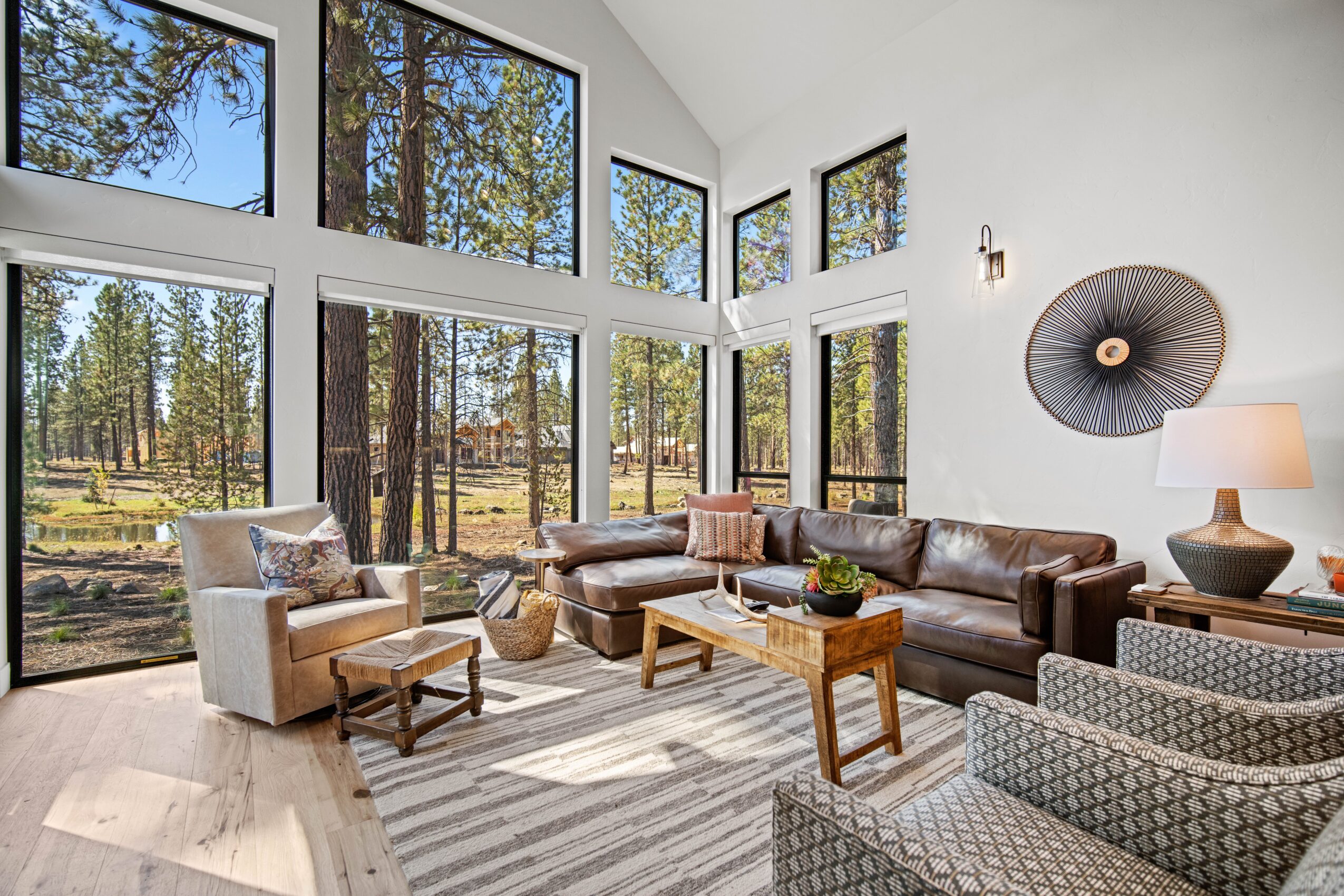 Bright living room with large windows showing trees outside, featuring a brown leather sofa, armchair, patterned chairs, and wooden coffee tables.
