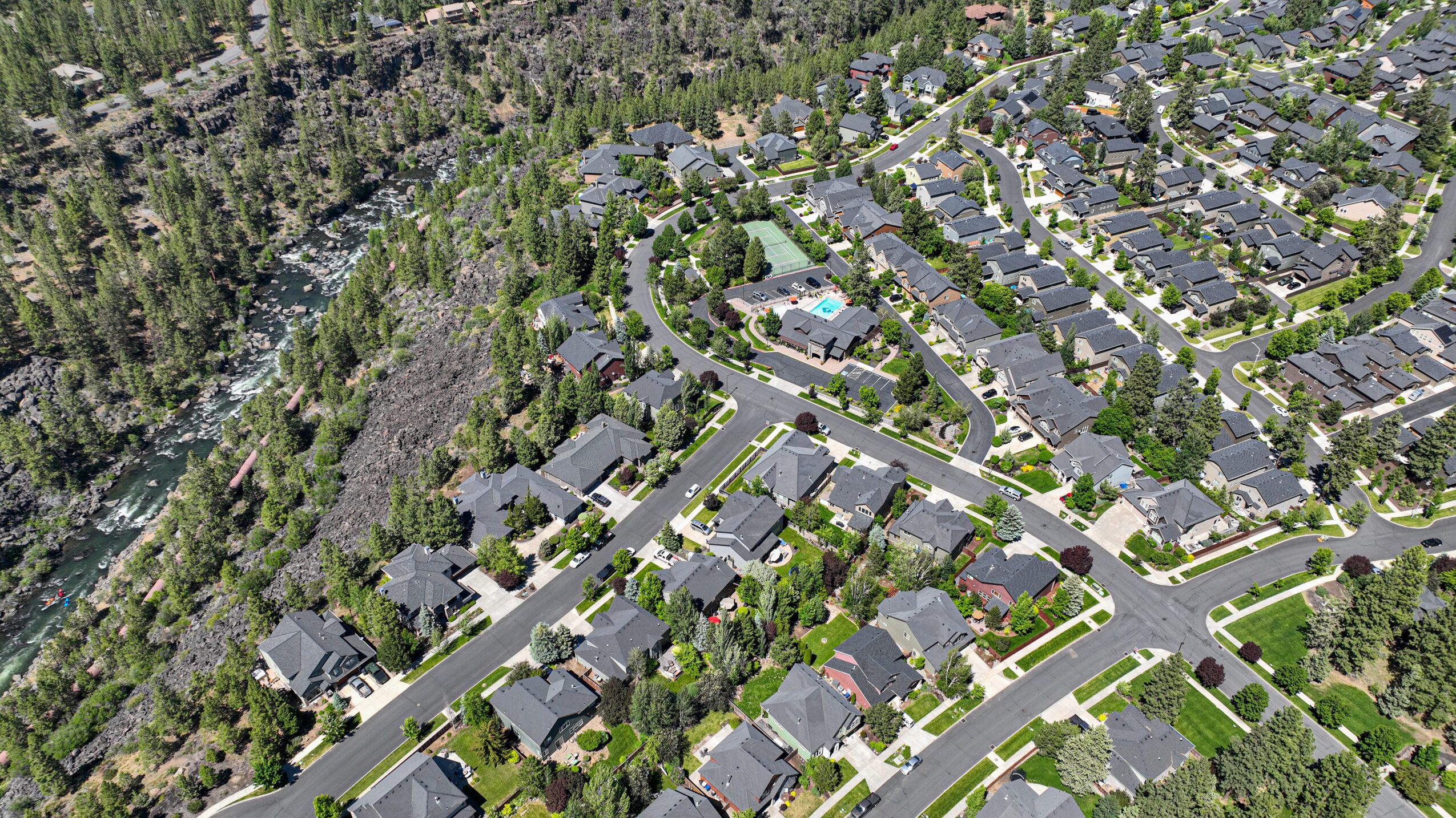 Aerial view of a suburban neighborhood with houses, streets, and a wooded area on the left side.