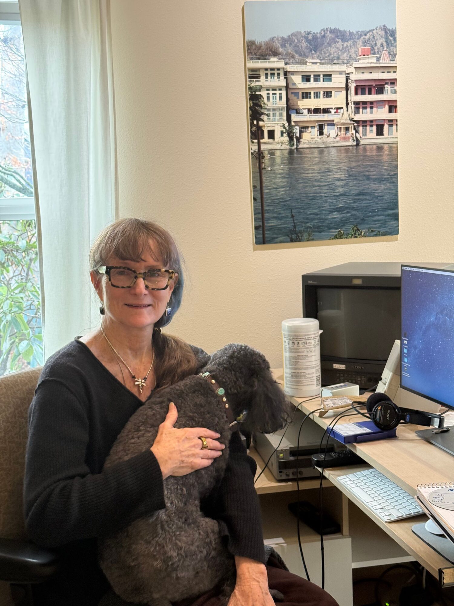 Woman holding a dog in an office, computer monitor and desk with items, window with curtains, and a painting of water and buildings.