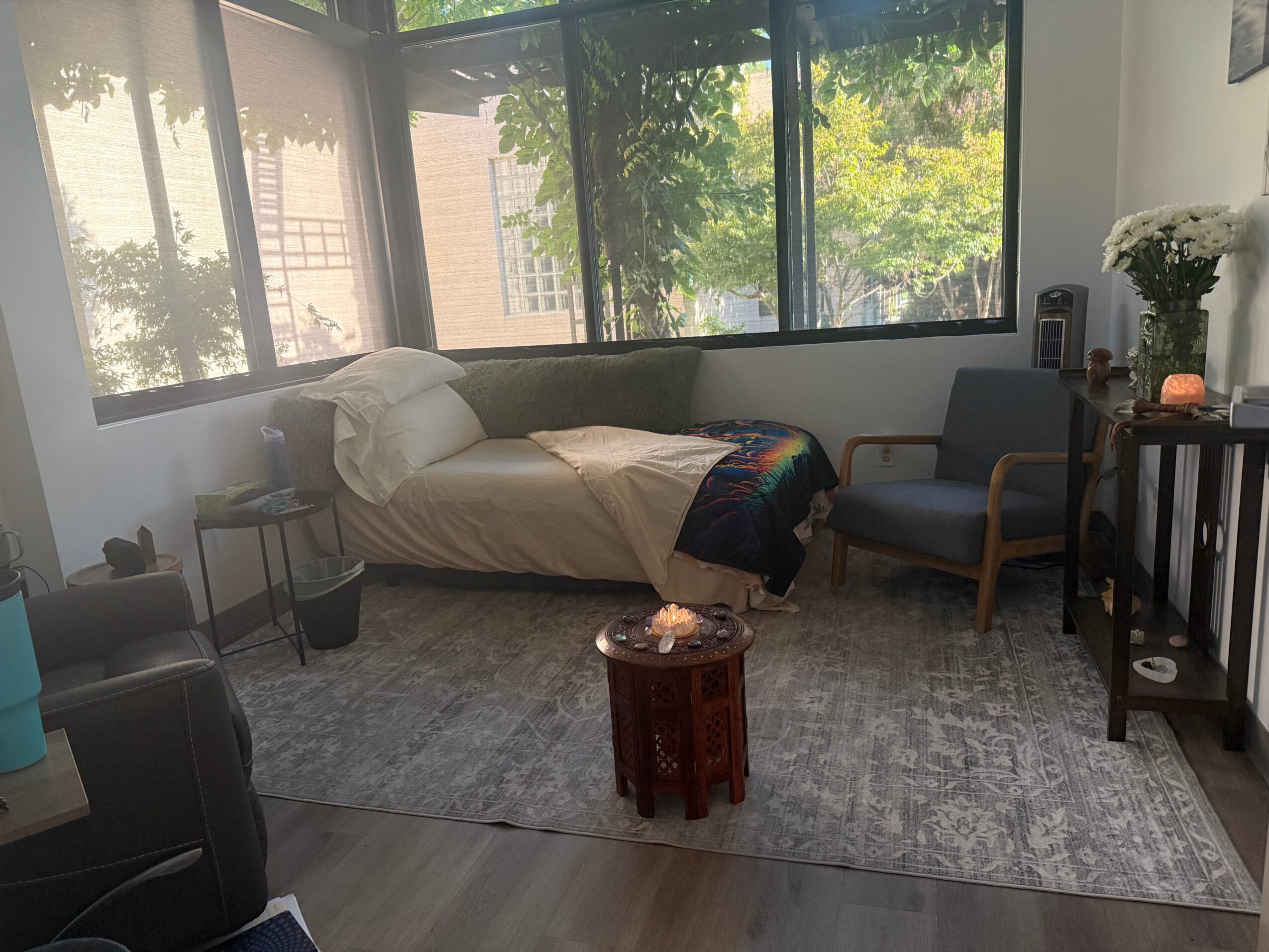 Bedroom with bed, armchair, small table, and large window with trees outside, sunlight streaming in.