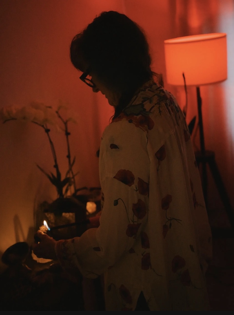 Person with glasses and curly hair in a dimly lit room with a lamp and plants, holding an object.