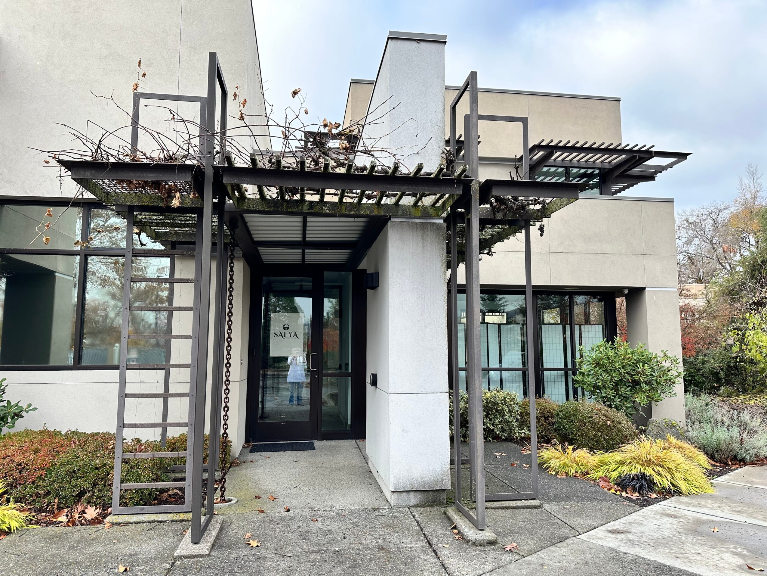 Modern building entrance with metal framework and plants, glass door, and surrounding landscaping.
