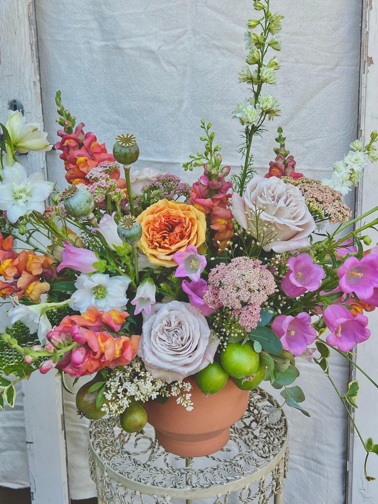 Colorful flower arrangement in a terracotta pot with green apples, on a decorative stand, against a light background.