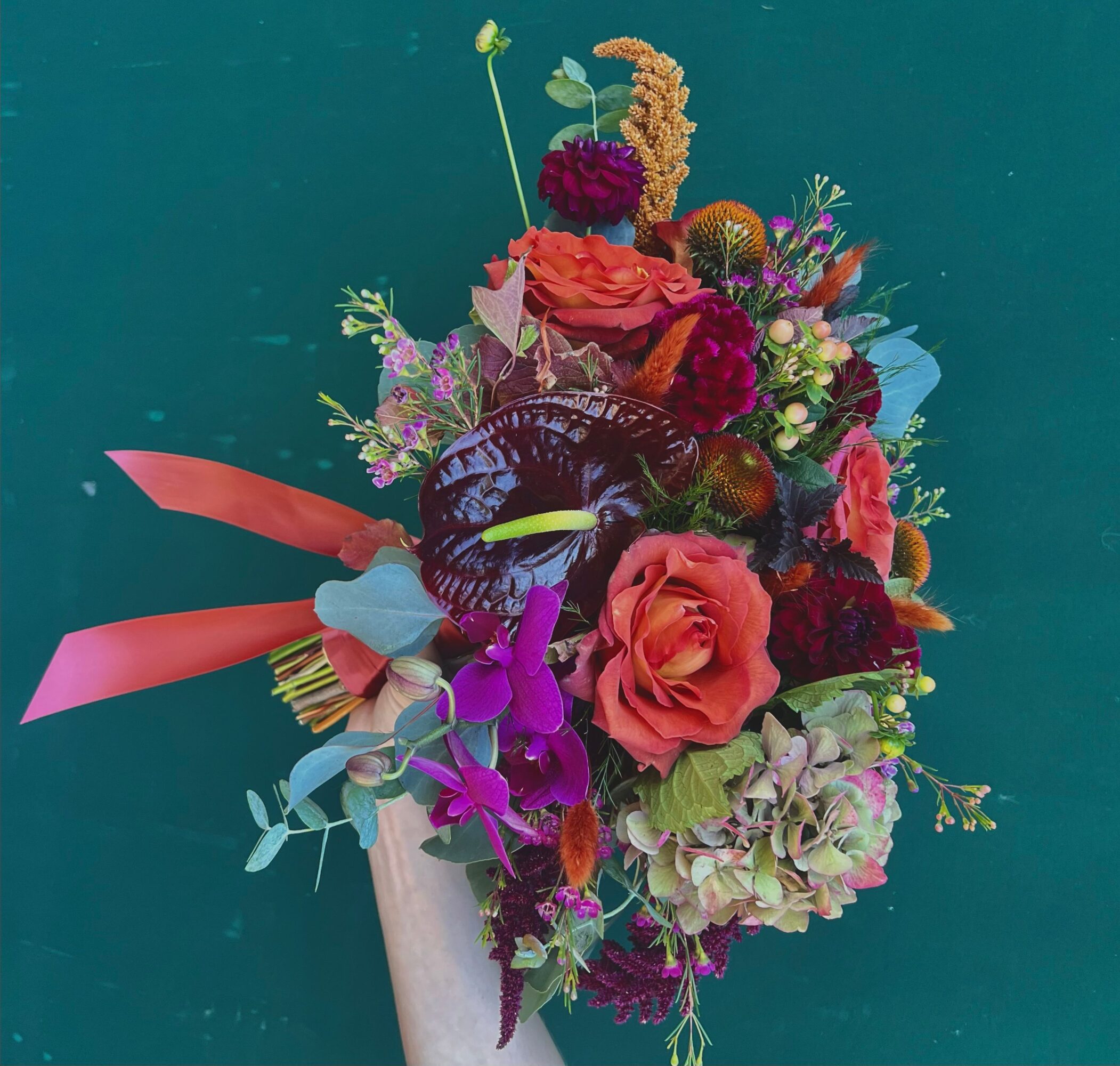 Colorful bouquet of flowers with pink, purple, and green blooms, held by a person against a teal background.