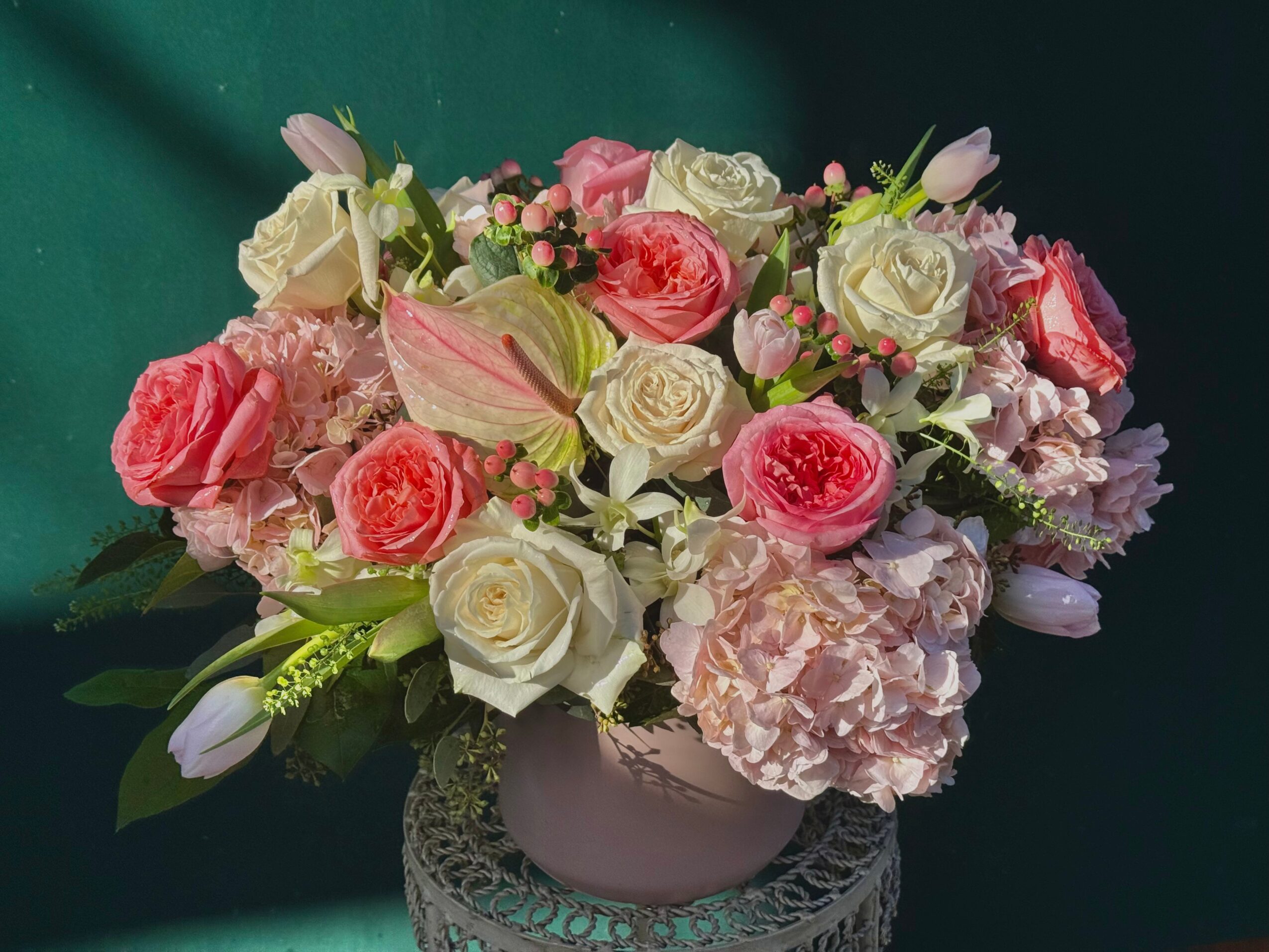 Flower arrangement with pink and white roses, hydrangeas, and tulips in a decorative vase, against a dark background.