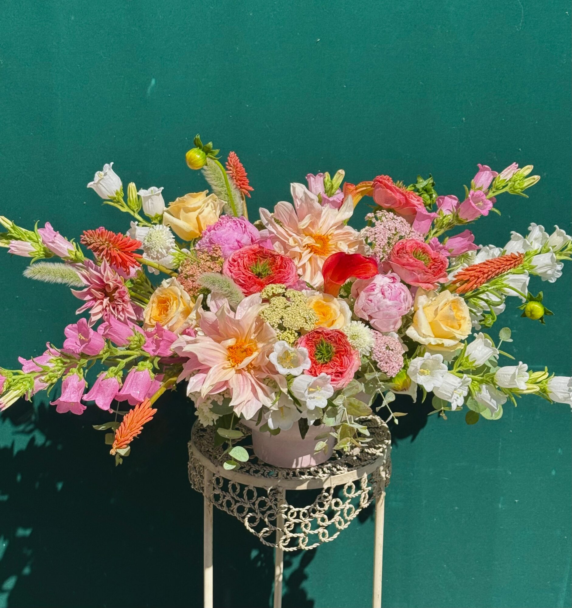 Colorful flower arrangement with pink, yellow, white, and red flowers in a white vase on a decorative stand against a green background.