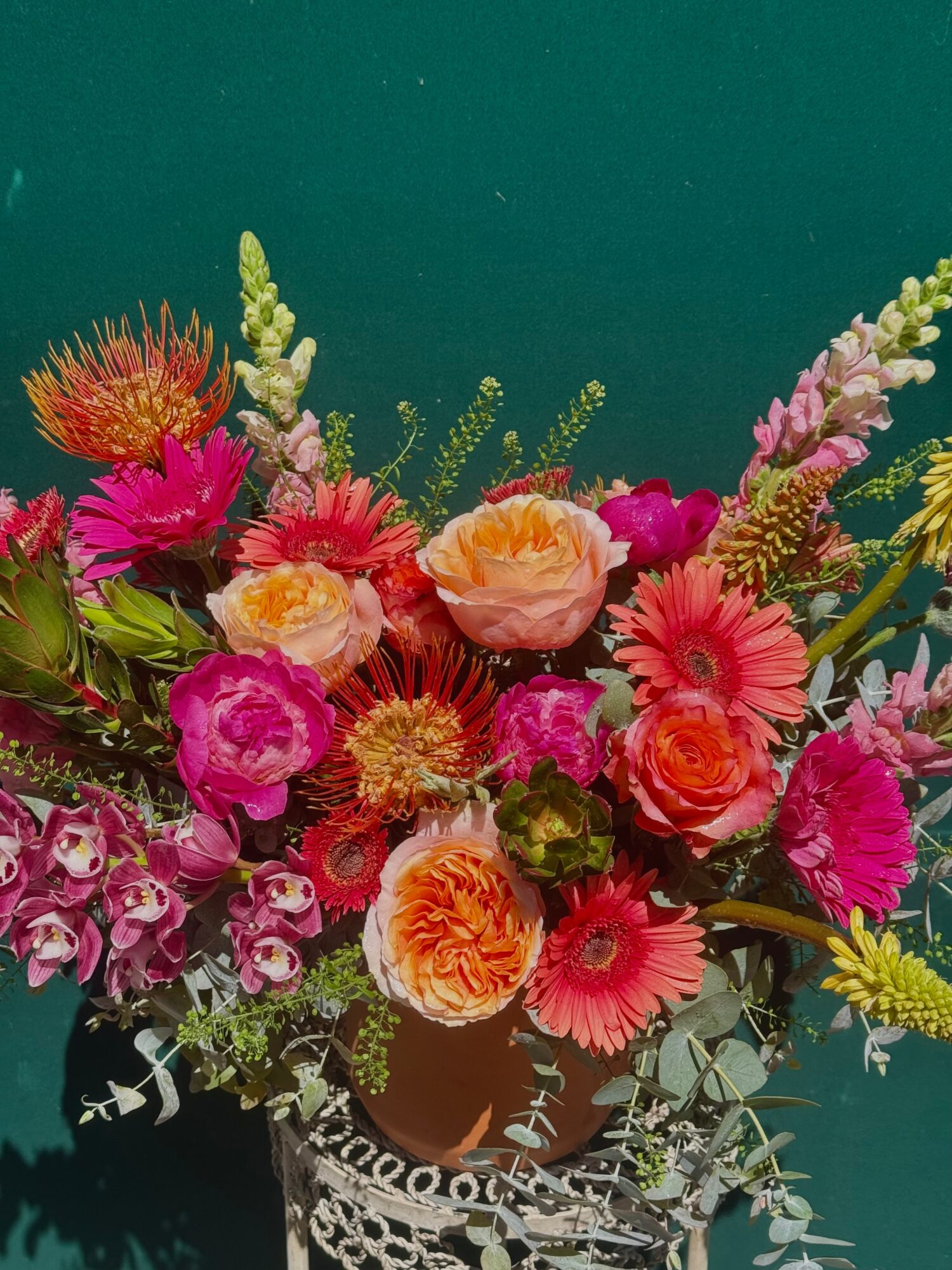 Colorful bouquet of various flowers in a pot, with pink, orange, and yellow blooms, against a teal background.