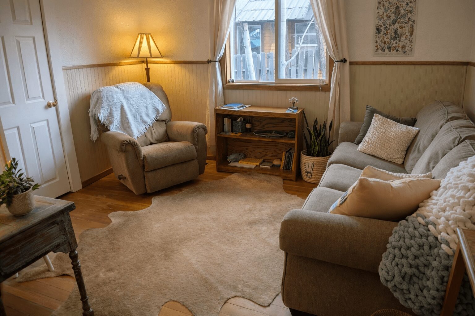 Living room with beige sofa, armchair, wooden side table, window, and warm lighting, carpeted floor, and decorative pillows.