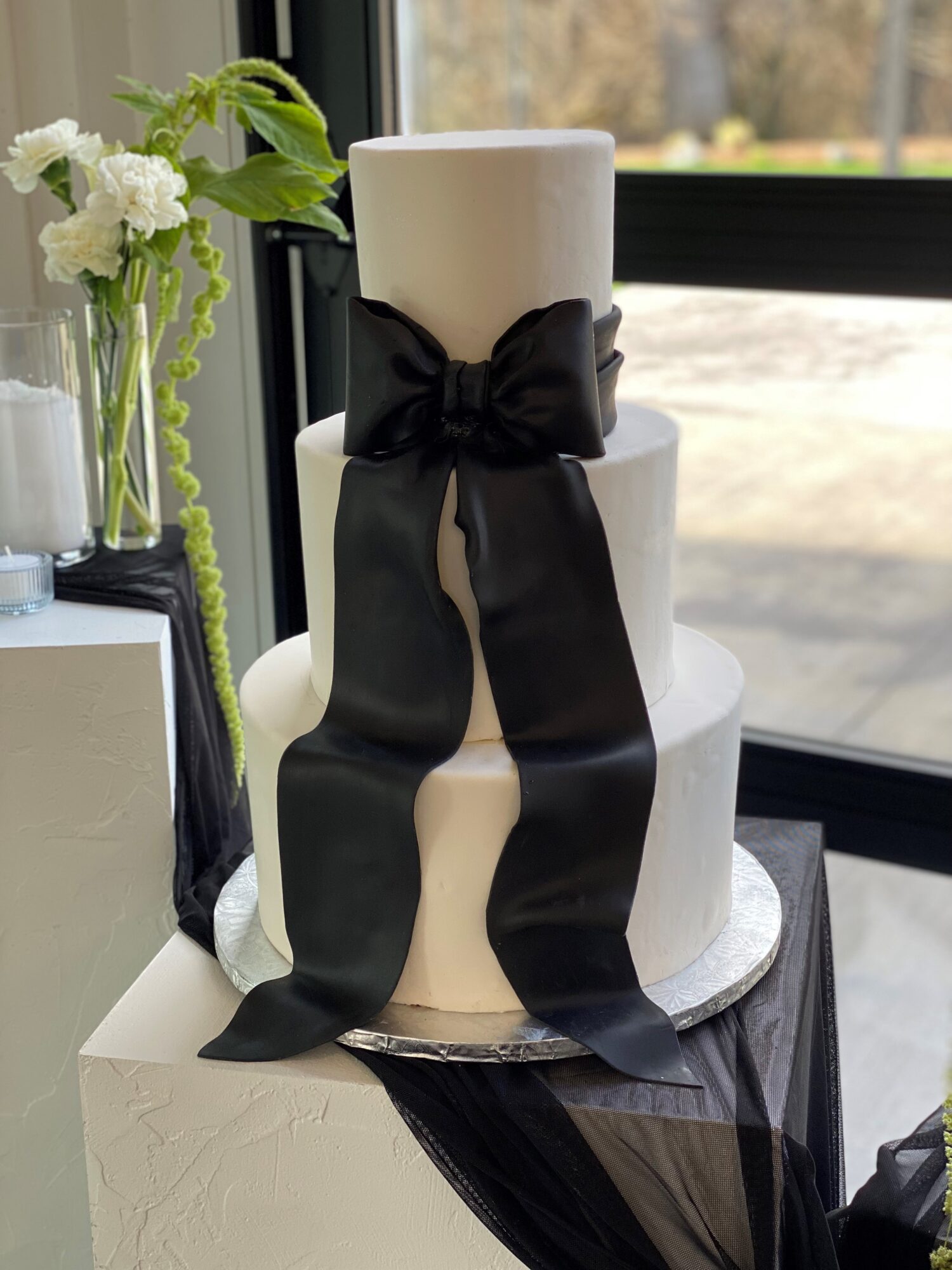 Three-tiered white cake with a large black ribbon bow, placed on a silver cake board near a window with flowers nearby.