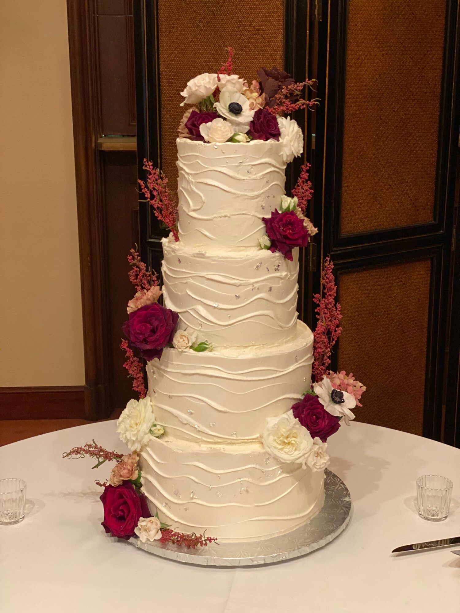 Tall white wedding cake with floral decorations on top and around base, placed on a table with a white tablecloth.