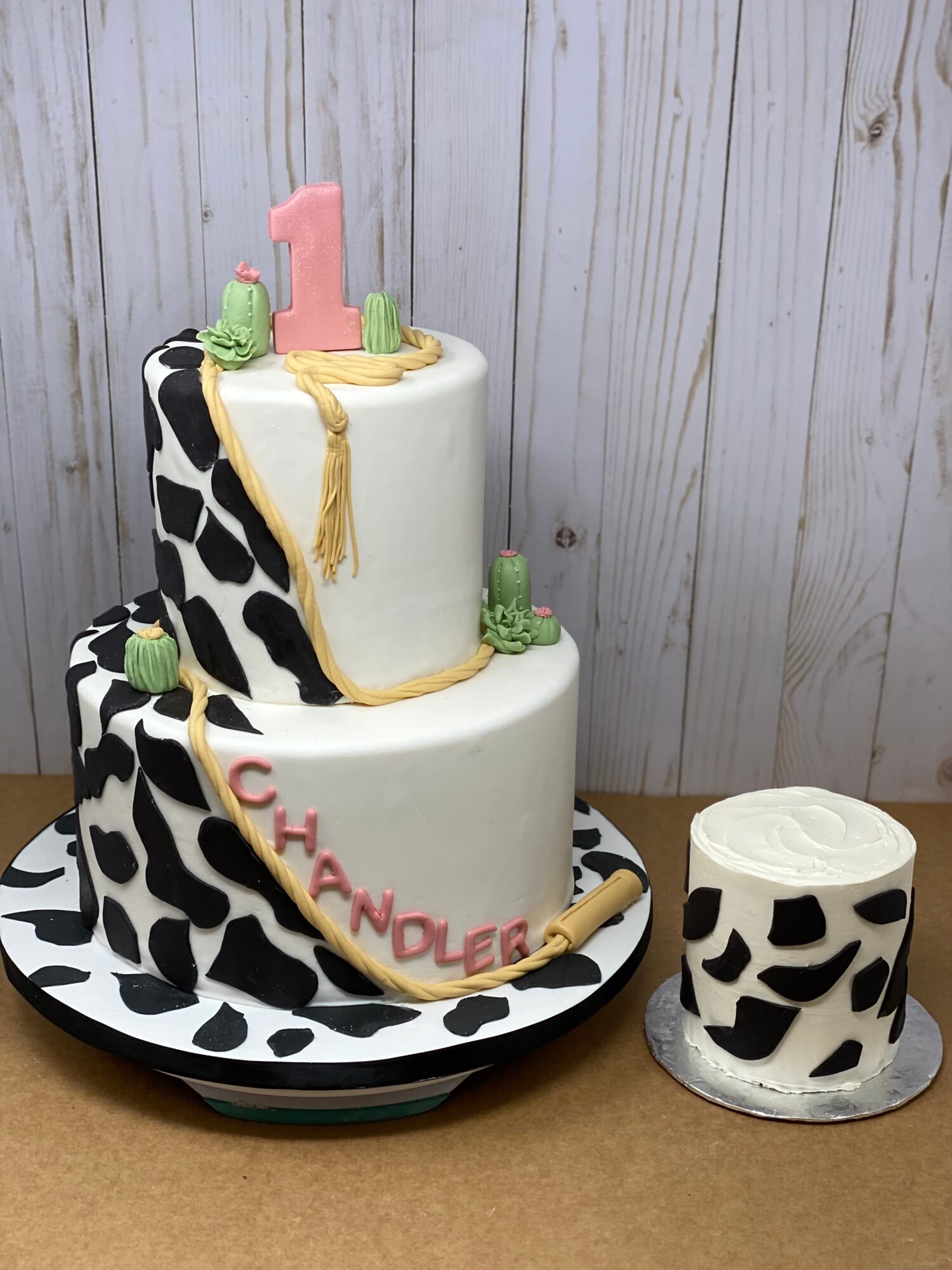 Two-tier cake with black and white cow print pattern, pink and green decorations, and a large pink number one on top.