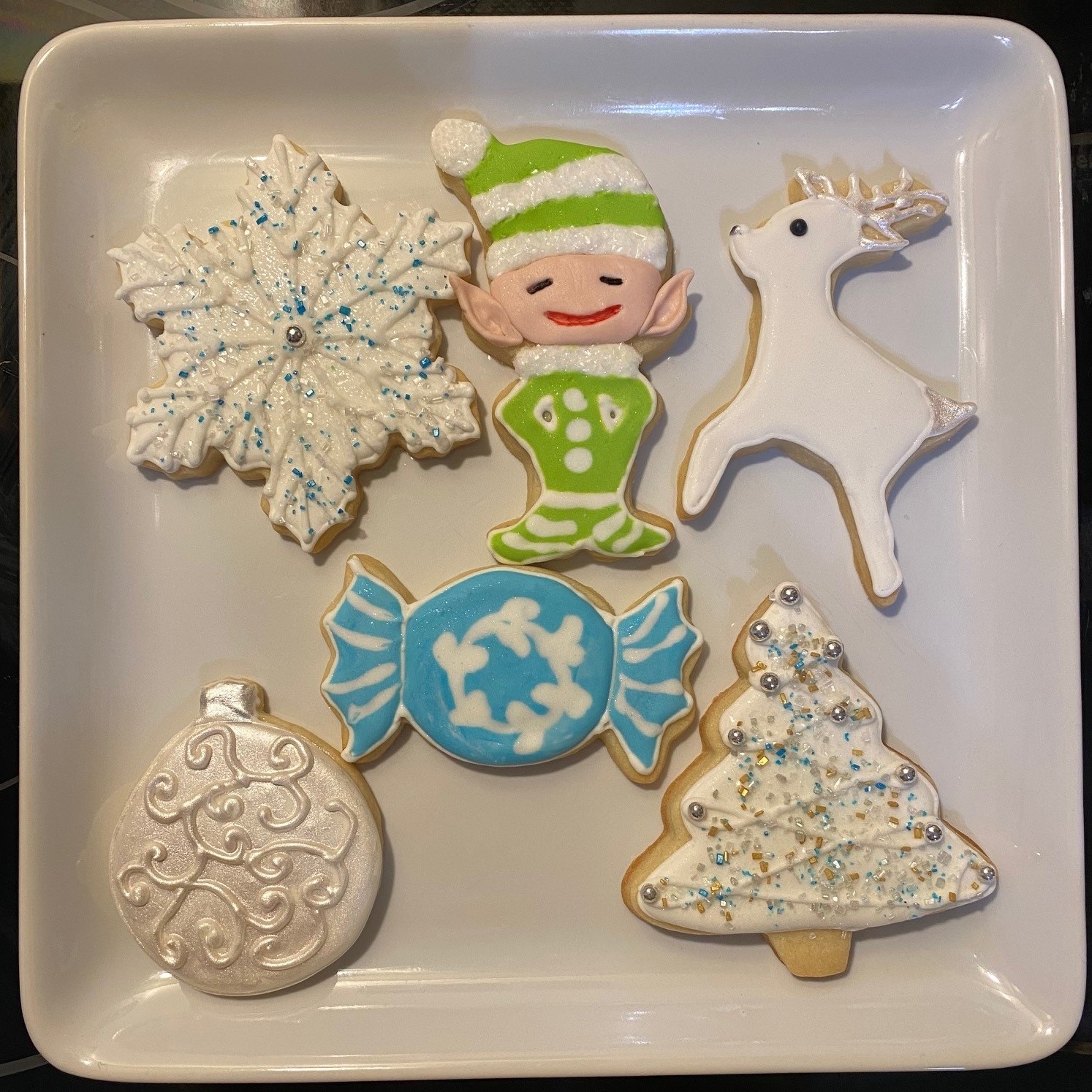 Plate with six decorated cookies: snowflake, elf, reindeer, candy, ornament, Christmas tree, all with icing and decorations.