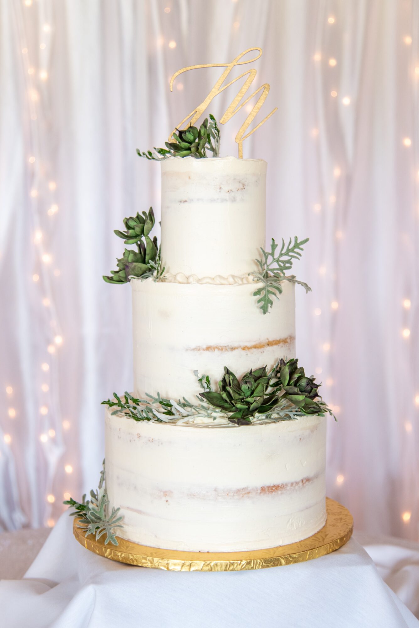 Three-tiered wedding cake decorated with greenery, set against a backdrop of white curtains and string lights.