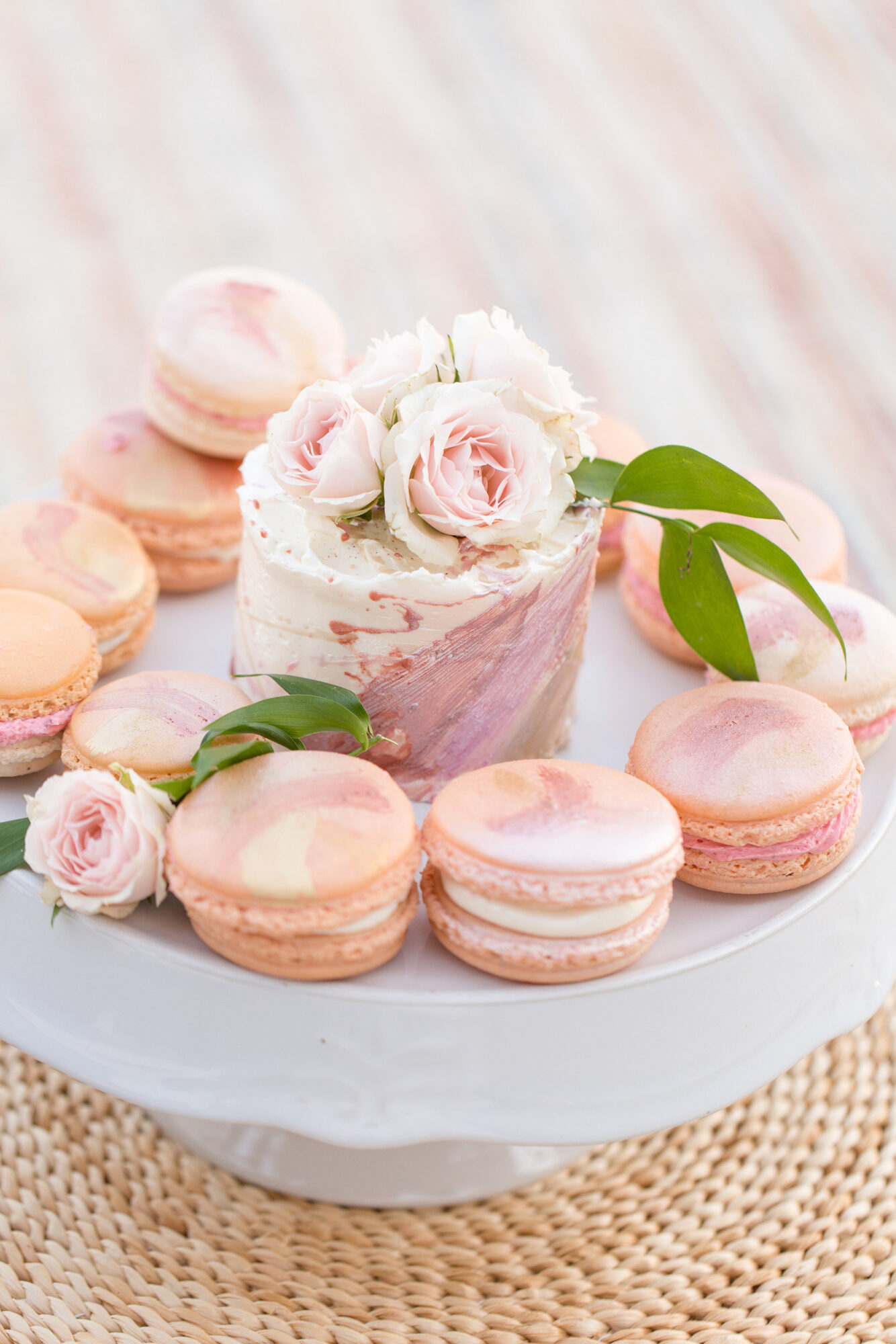 Cake decorated with pink roses and macarons on a white cake stand, with green leaves, on a woven surface.