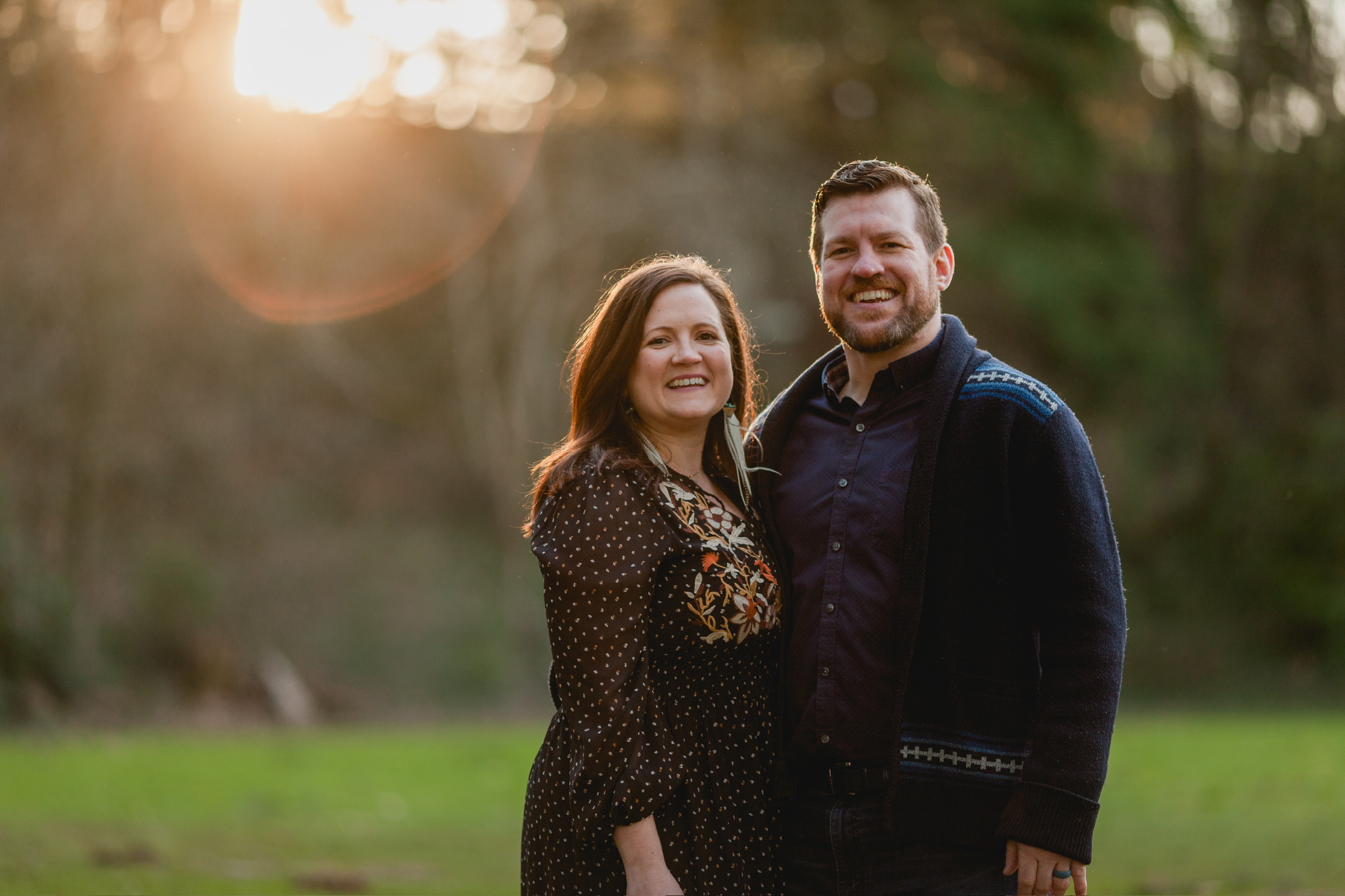 A smiling couple standing outdoors in a park during sunset, with trees in the background.