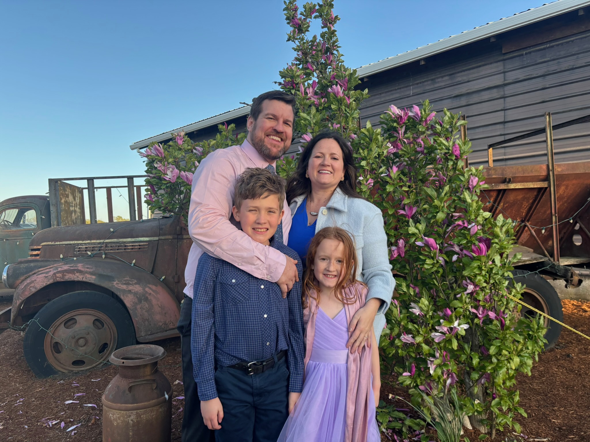 A family of four stands outdoors near a flowering bush, smiling, with a vintage truck and building in background.